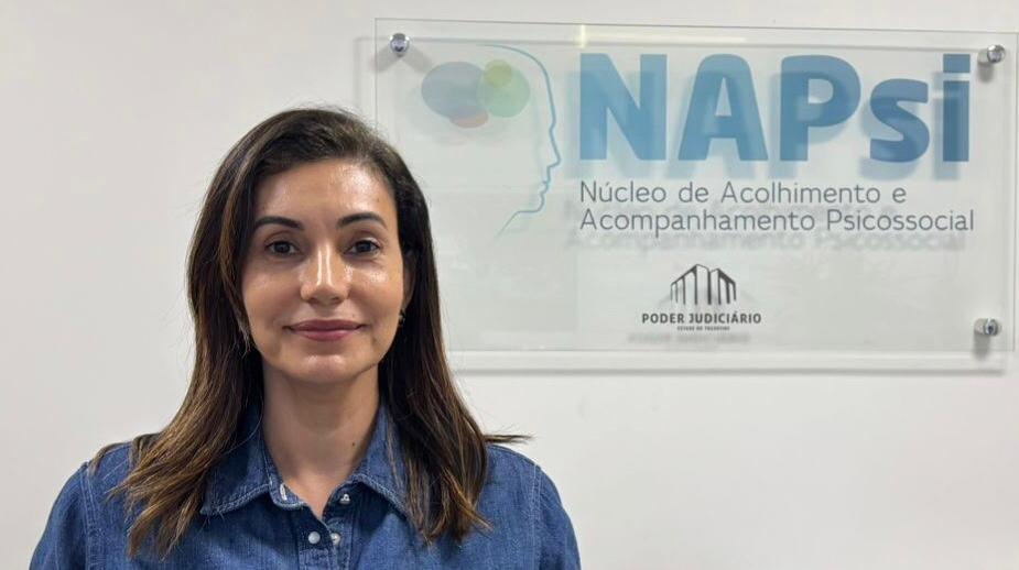A foto mostra uma mulher de cabelos castanhos lisos, na altura dos ombros, vestindo uma camisa jeans. Ela está posicionada à frente de uma parede branca, onde há uma placa institucional com o logotipo do NAPsi – Núcleo de Acolhimento e Acompanhamento Psicossocial, do Poder Judiciário do Estado do Tocantins.