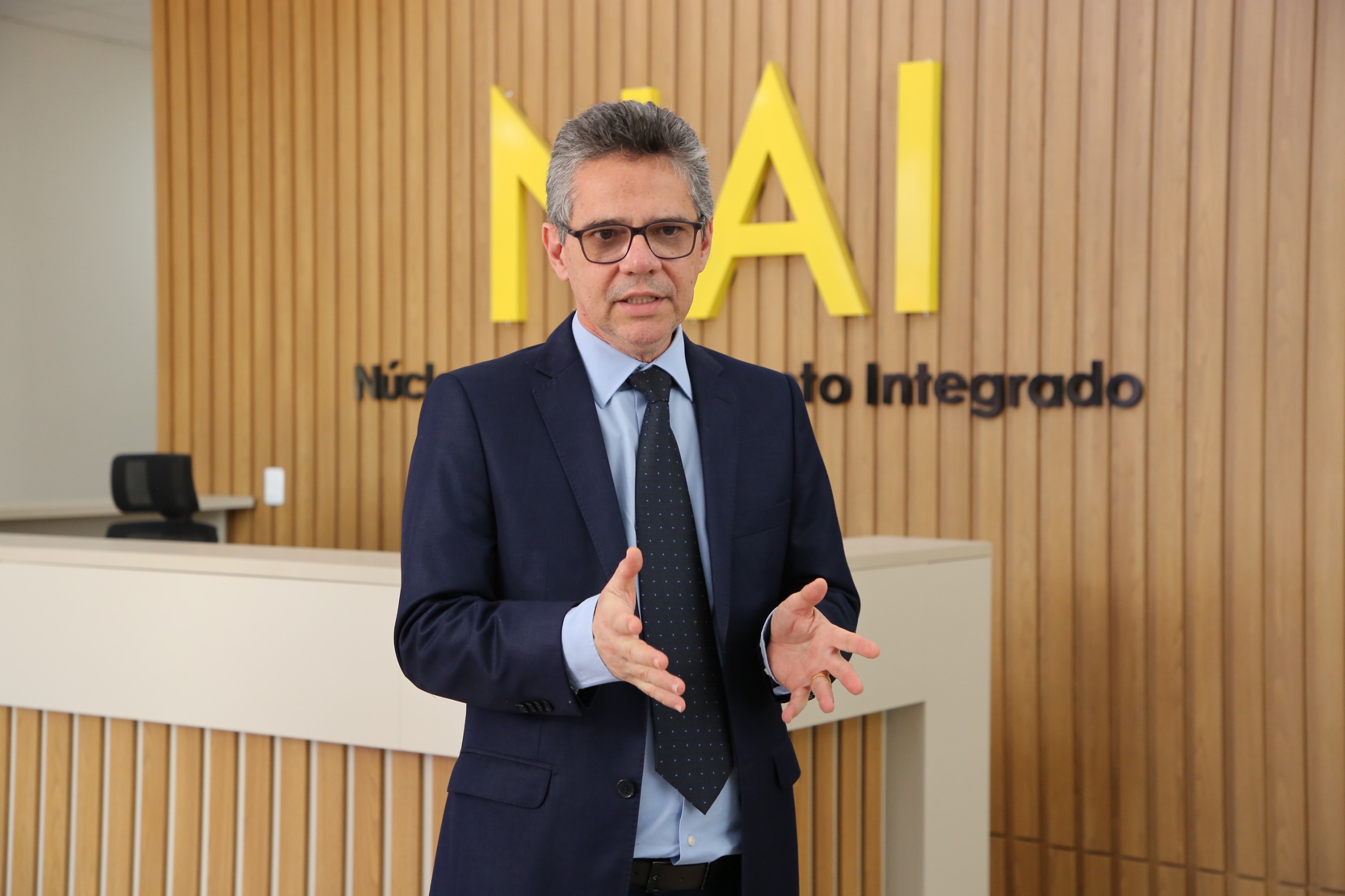 Imagem mostra o juiz Adriano Gomes,  de terno azul-escuro e gravata, gesticulando enquanto fala, em um ambiente institucional. Ao fundo, há uma parede com revestimento em madeira clara e a sigla “NAI” em letras grandes e amarelas, que se refere ao Núcleo de Atendimento Integrado.