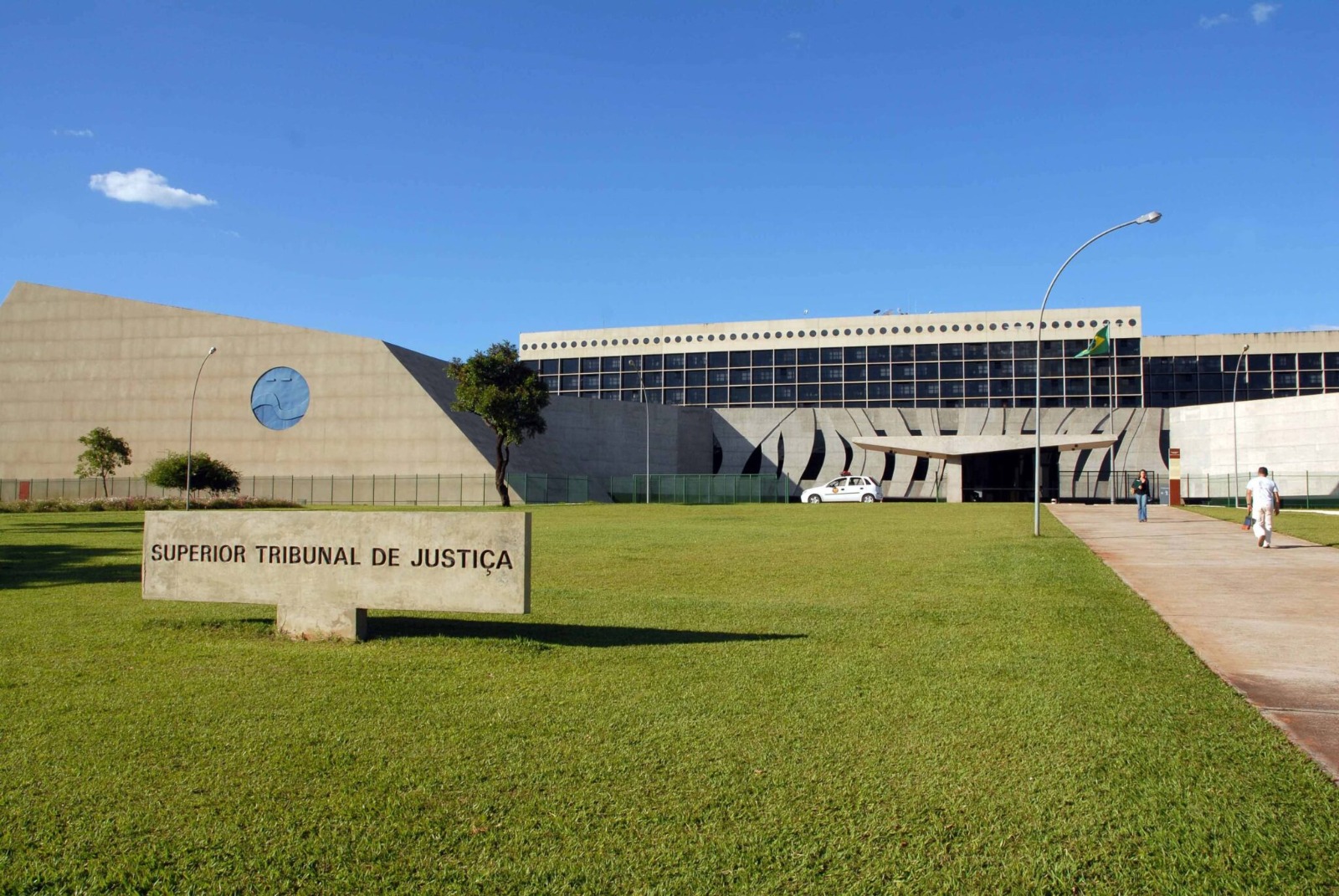 A imagem mostra a fachada moderna do edifício do Superior Tribunal de Justiça (STJ), em Brasília, sob um céu azul e ensolarado. O prédio tem linhas geométricas marcantes em concreto aparente e janelas escuras em sua parte superior. No gramado à frente, há uma placa de concreto com a inscrição “SUPERIOR TRIBUNAL DE JUSTIÇA” em letras pretas.