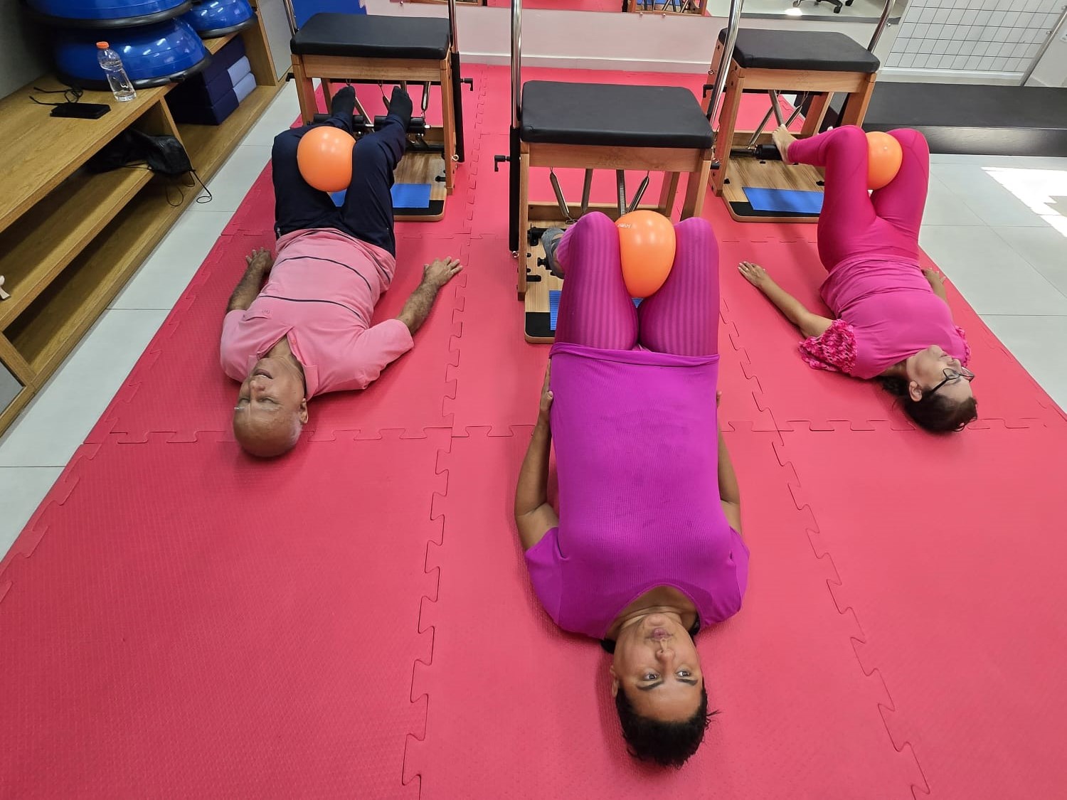 A imagem mostra três pessoas deitadas em esteiras vermelhas, vestidas de rosa, realizando exercícios com bolas laranjas em um estúdio de Pilates, em alusão ao Outubro Rosa — mês dedicado à prevenção do câncer de mama e do colo do útero.