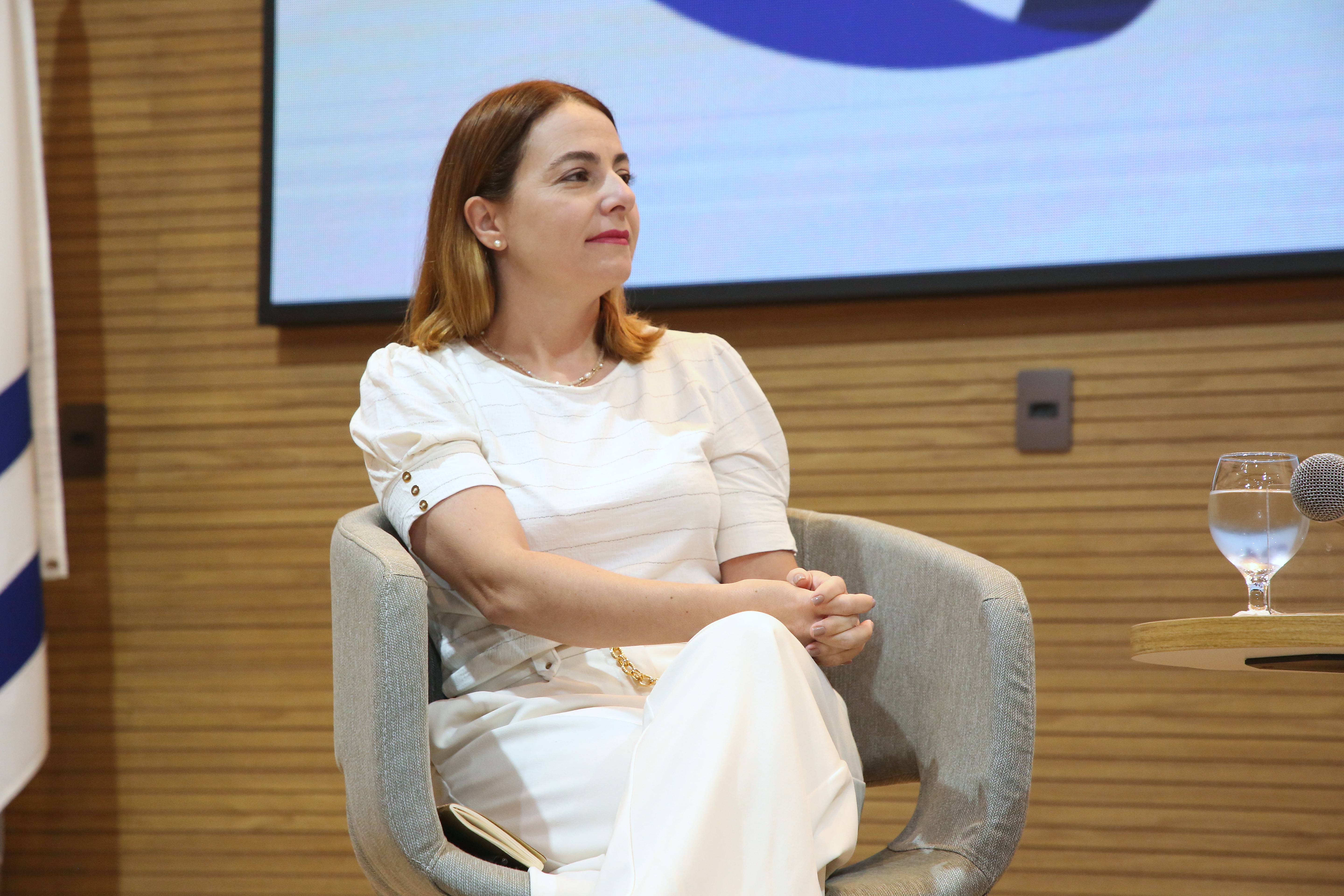 Cristiana é uma mulher branca de cabelos claros e usa uma roupa branca; ela está sentada no palco do auditório, durante palestra