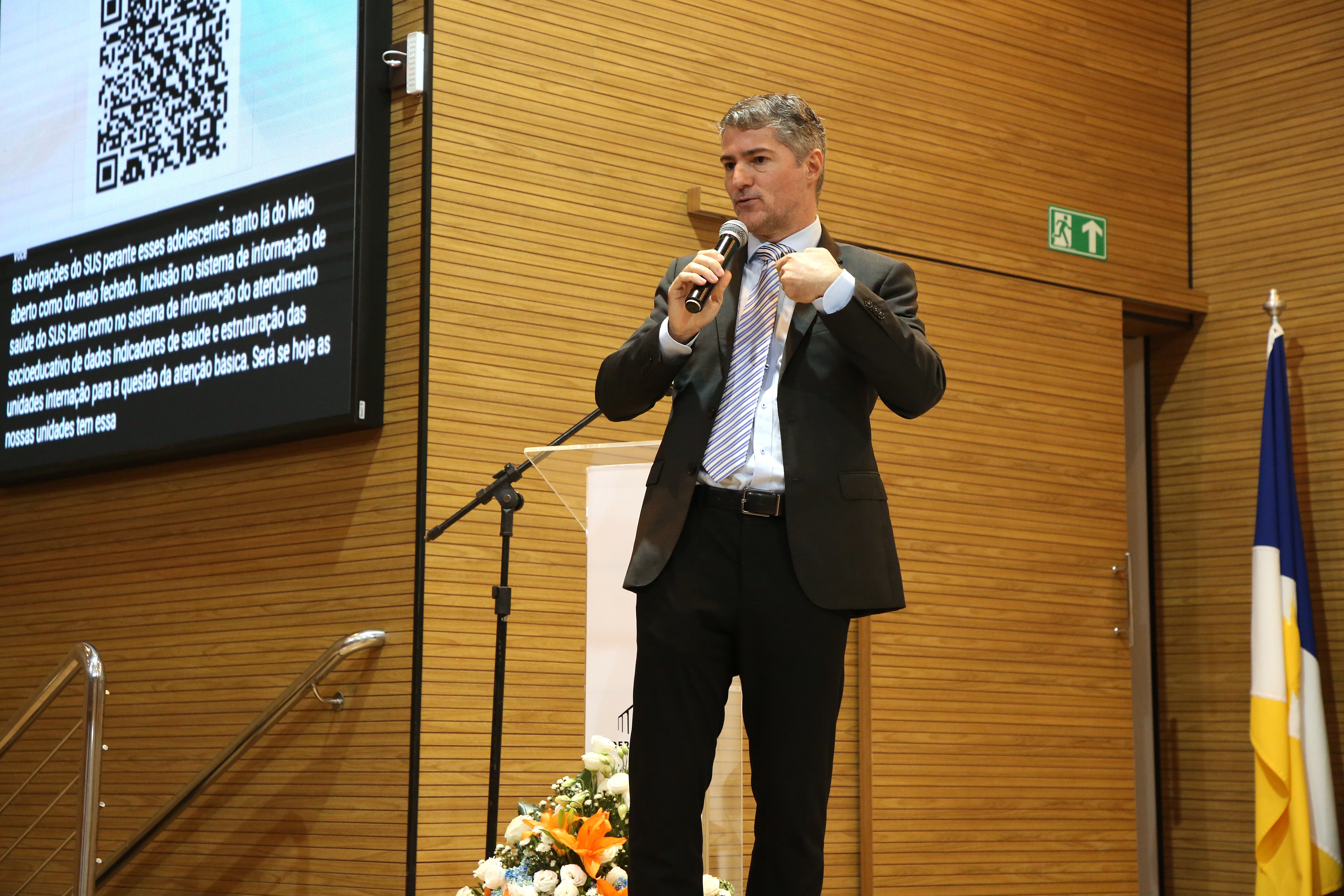 Promotor de Justiça Sidney Fiore, um homem branco de cabelos grisalhos, fala ao microfone durante palestra; ele usa terno escuro e camisa branca 