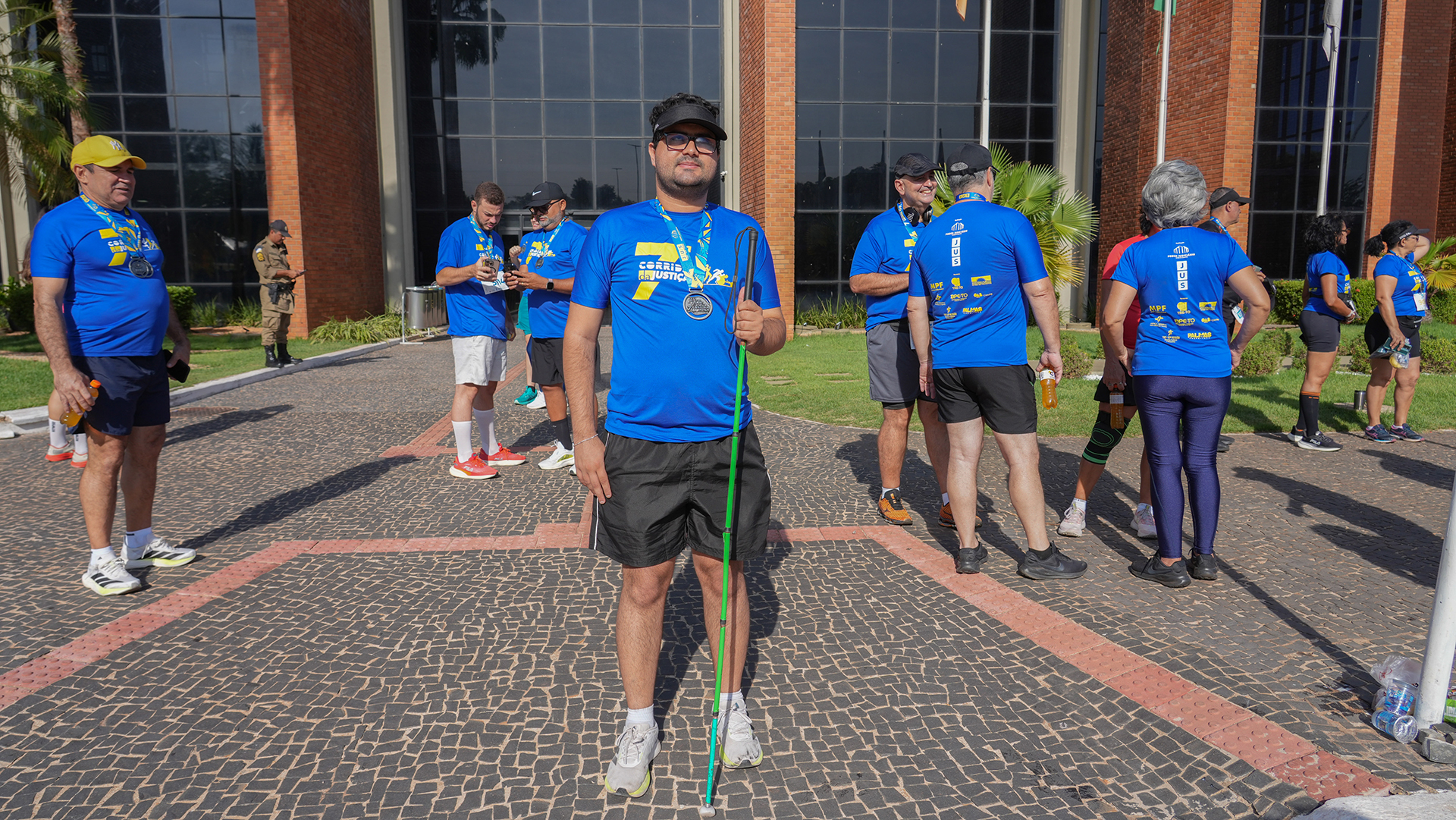 Rapaz com baixa visão participante da corrida; ele está em pé em frente ao prédio do TJTO e usa bermuda preta e camiseta azul da corrida e um guia