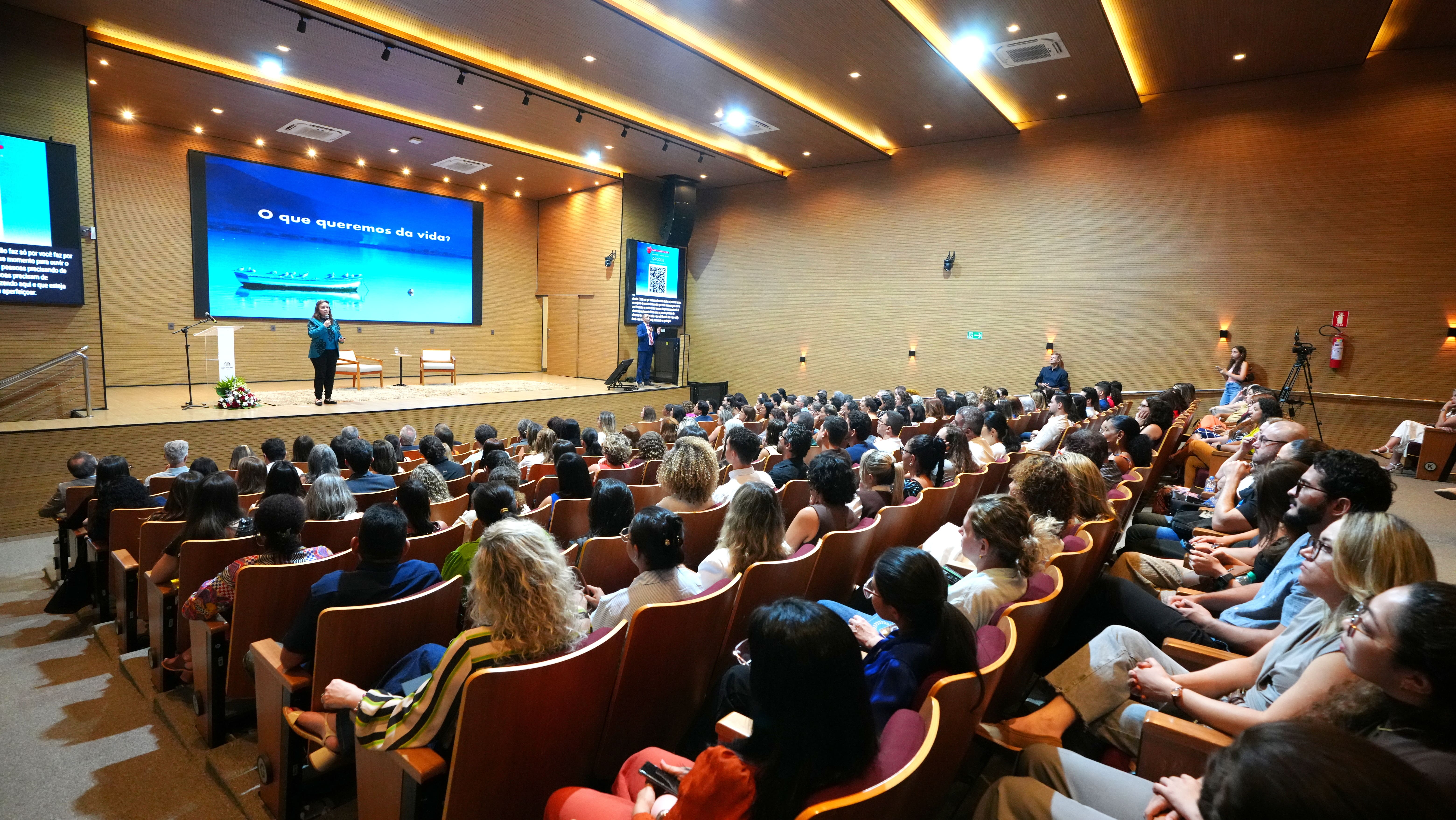 A imagem mostra o auditório do TJTO lotado, com pessoas atentamente assistindo à palestra da professora e filósofa Lúcia Helena Galvão, que está no palco e deu início à X Semana da Saúde do Poder Judiciário Tocantinense. No telão atrás dela, aparece a pergunta: "O que queremos da vida?".