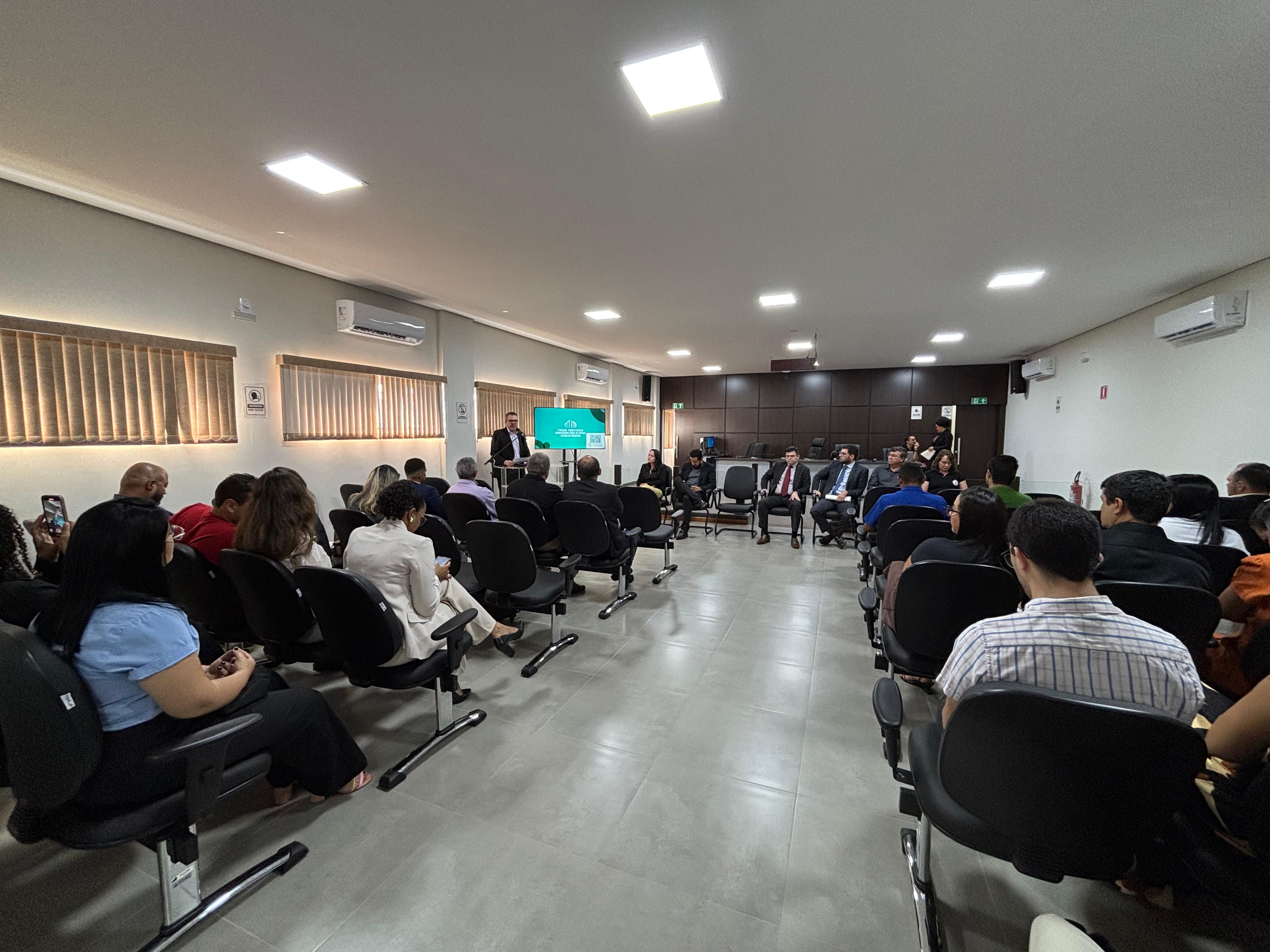 A imagem mostra o Salão do Júri durante as correições gerais ordinárias. Fileiras de cadeiras estão alinhadas dos dois lados de um corredor central. À frente, um grupo de autoridades está sentado enquanto um juiz fala ao púlpito.