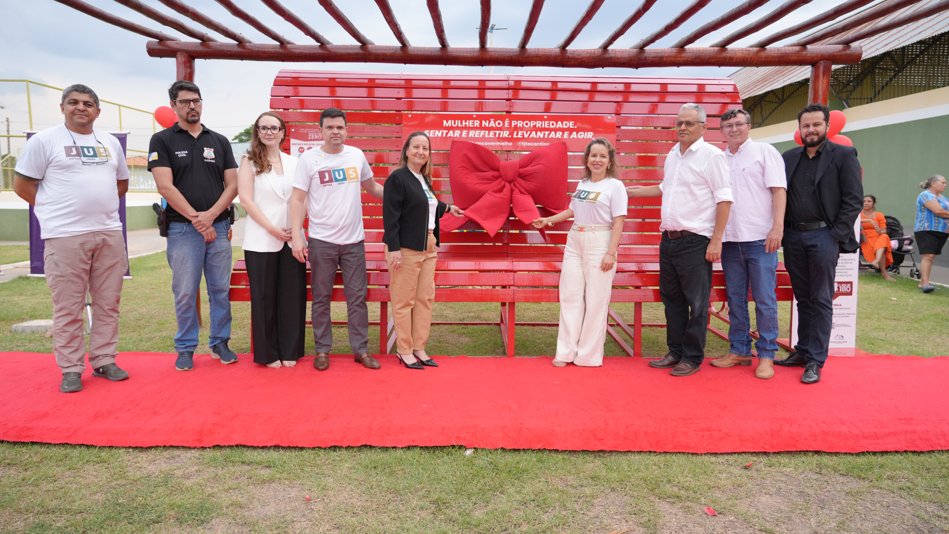 Em um espaço ao ar livre, sobre um tapete vermelho, nove pessoas posam para uma foto em frente a um Banco Vermelho, instalado sob uma estrutura de madeira. O banco traz uma grande fita vermelha em formato de laço e uma placa com os dizeres: “Mulher não é propriedade. Sentar e refletir. Levantar e agir.” O grupo é composto por homens e mulheres — alguns vestindo camisetas brancas do projeto “JUS” e outros com trajes sociais ou camisas de trabalho. O clima é de solenidade e união na luta contra a violência de gênero.