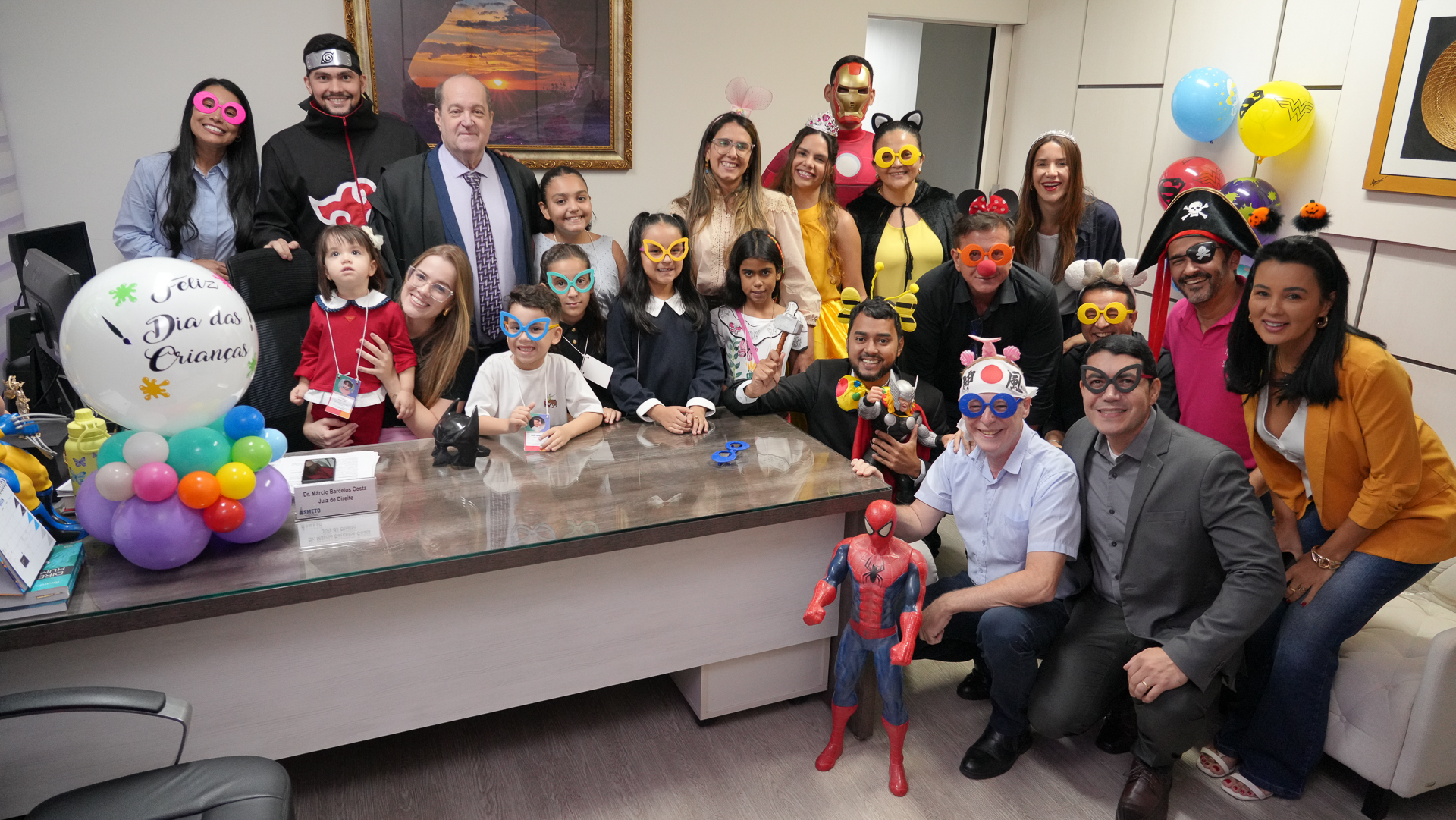 Grupo de adultos e crianças fantasiados e usando adereços coloridos posam sorrindo em um escritório decorado com balões, durante ação comemorativa pelo Dia das Crianças no Tribunal de Justiça do Tocantins.