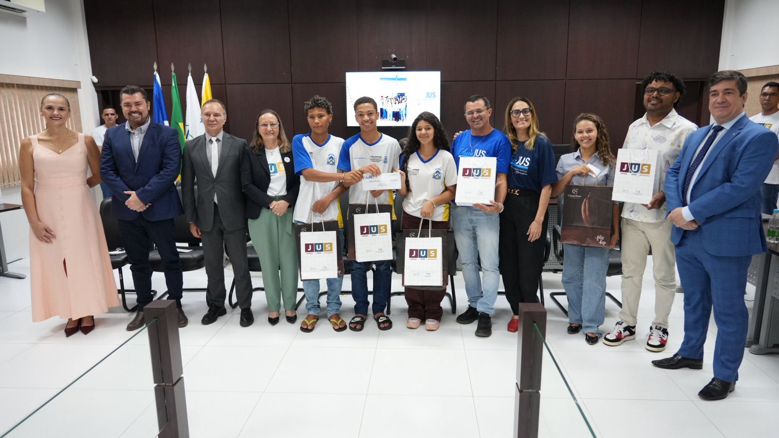 A imagem mostra jovens com sacolas nas mãos durante a premiação do projeto EducaJus, em Novo Acordo, em uma cerimônia formal com autoridades e organizadores.