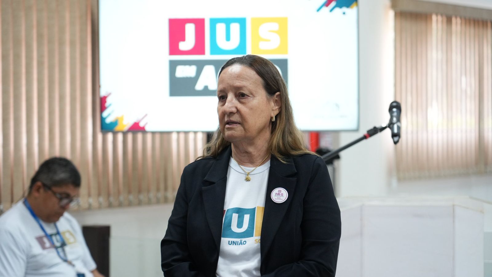 A imagem mostra a presidente do TJTO em primeiro plano, com uma pessoa sentada ao fundo e um telão exibindo a logo do projeto “JUS em Ação”.