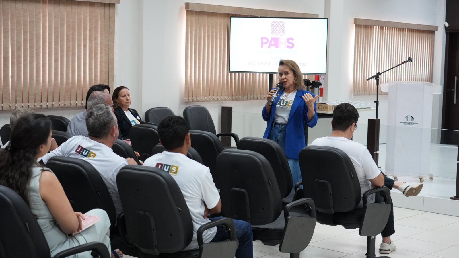Foto aberta de um auditório com pessoas assistindo palestra da juíza Cirlene, que está ao fundo usando roupa azul e uma camiseta branca