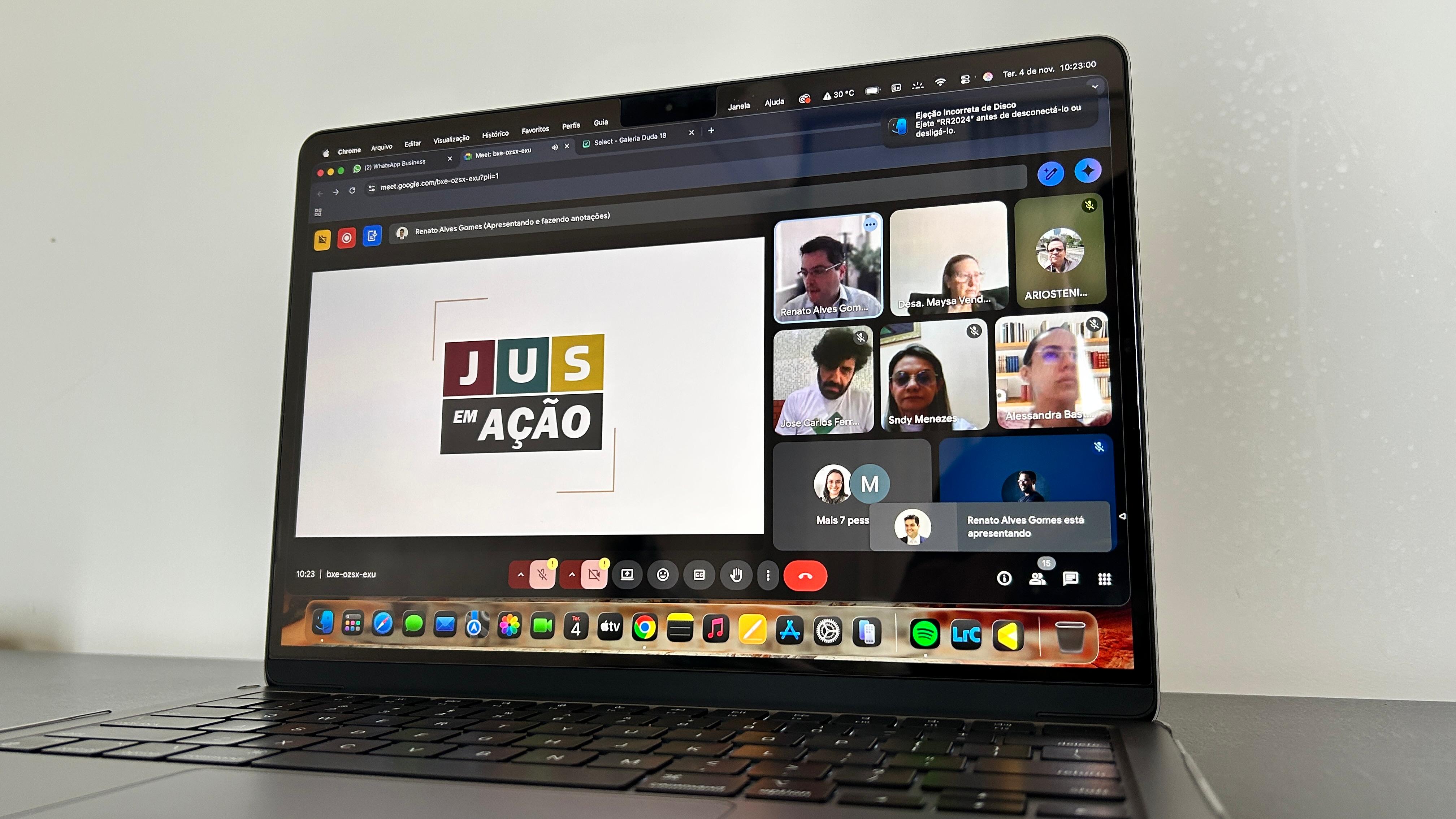 A imagem mostra a tela de um laptop aberto, exibindo uma videochamada com várias pessoas, com o logotipo "Jus em Ação" em destaque na tela.