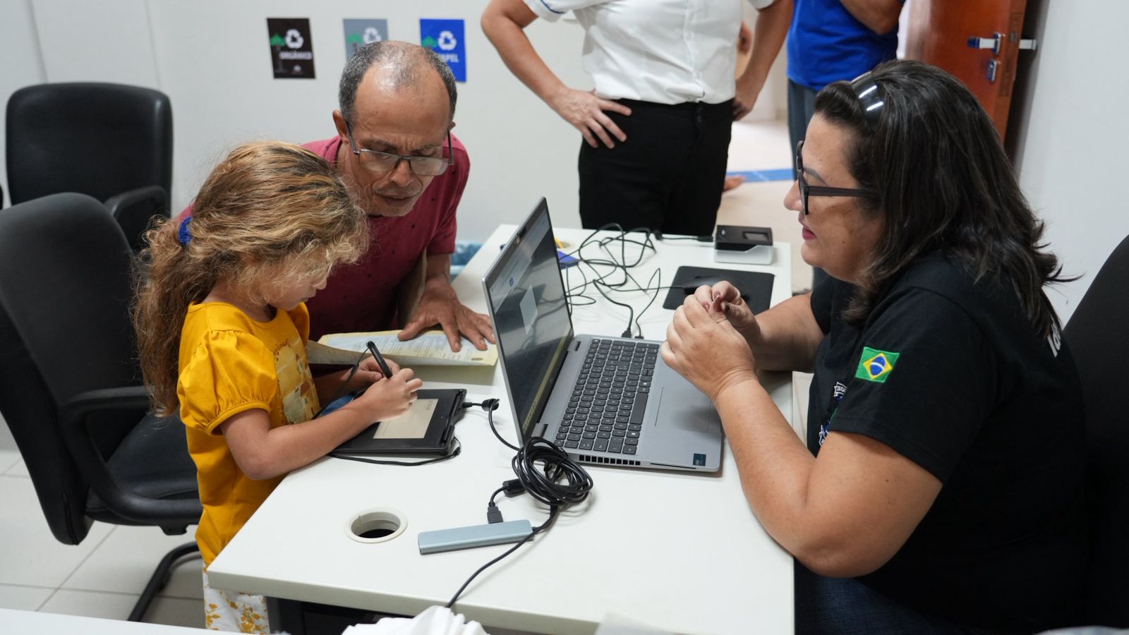 A imagem mostra um momento de interação durante o projeto JUS em Ação, em Novo Acordo. Uma criança, auxiliada por adultos, assina um documento digital.
