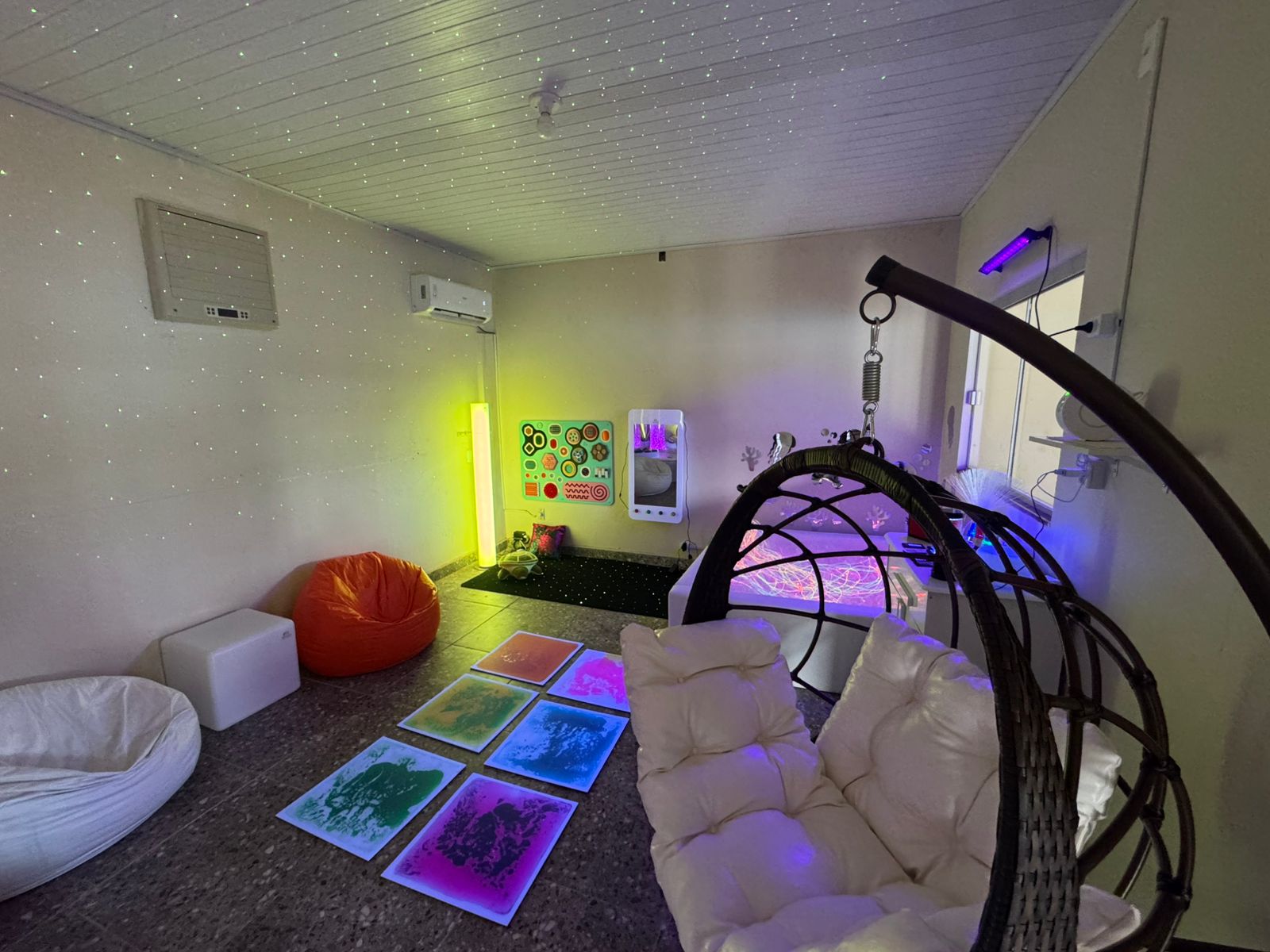 A sala apresenta um ambiente de estimulação sensorial, com iluminação colorida suave e pontos luminosos espalhados pelo teto e paredes. Há pufes, poltronas e tapetes interativos no chão, além de painéis sensoriais na parede. No canto direito, uma cadeira suspensa acolchoada compõe o espaço, que também conta com equipamentos de luz e fibras óticas usados para atividades terapêuticas.