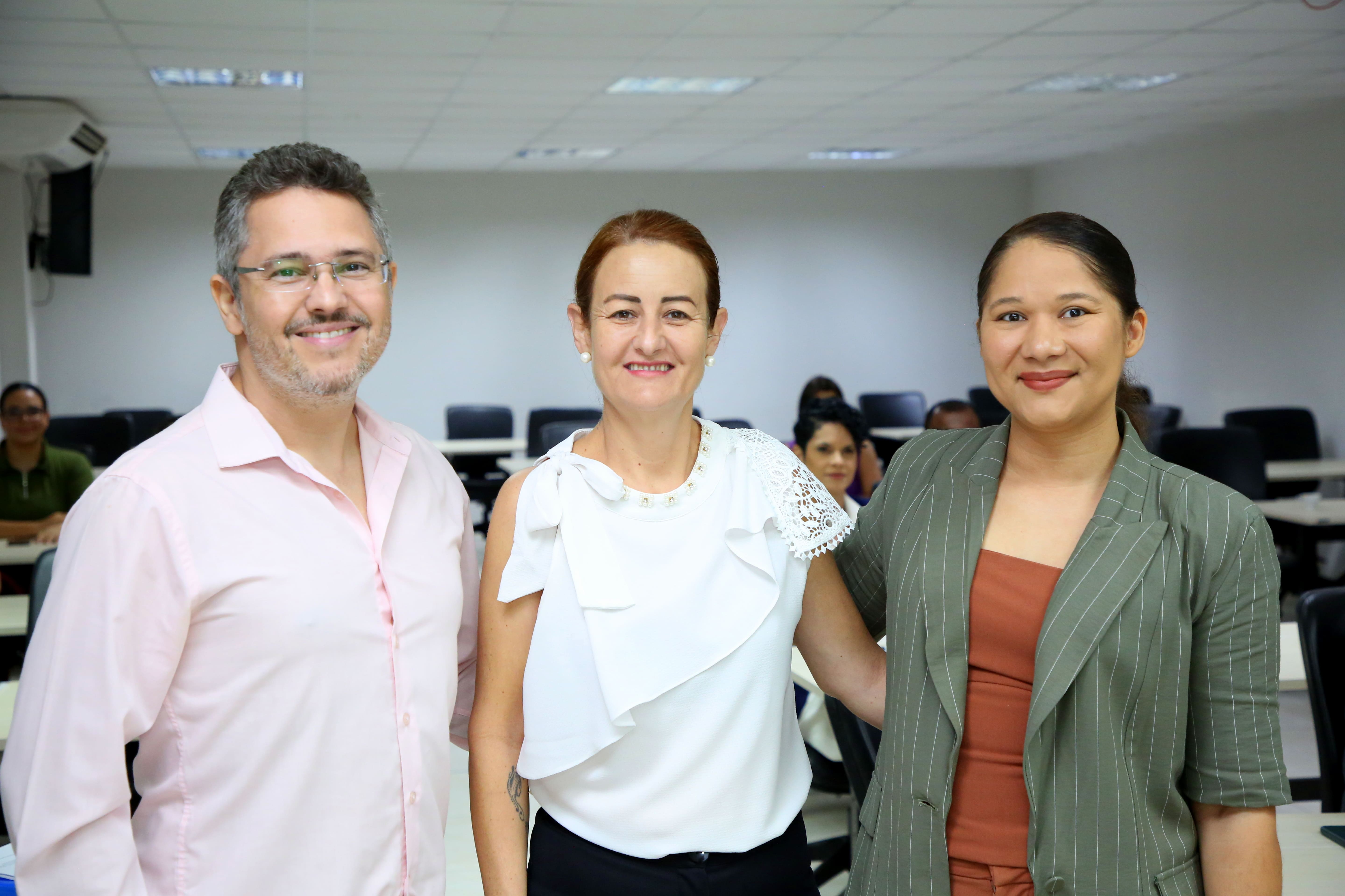 Três pessoas estão em pé, sorrindo para a câmera, em um ambiente interno de sala de reunião ou treinamento. Ao fundo, é possível ver mesas, cadeiras e outras pessoas sentadas. A foto transmite clima de cordialidade e profissionalismo.