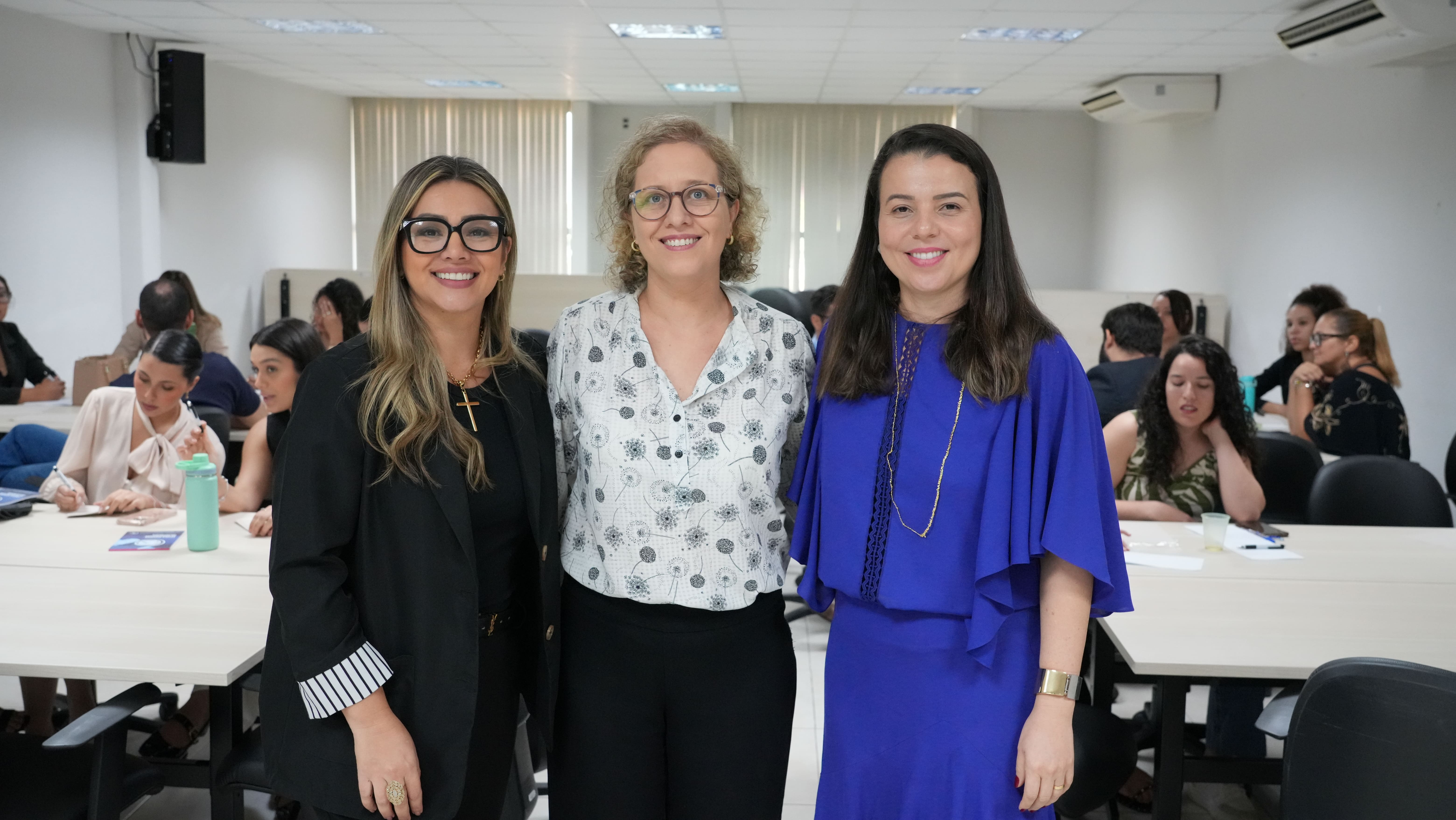 Três mulheres posam sorridentes em sala de aula durante atividade em grupo, com participantes ao fundo.
