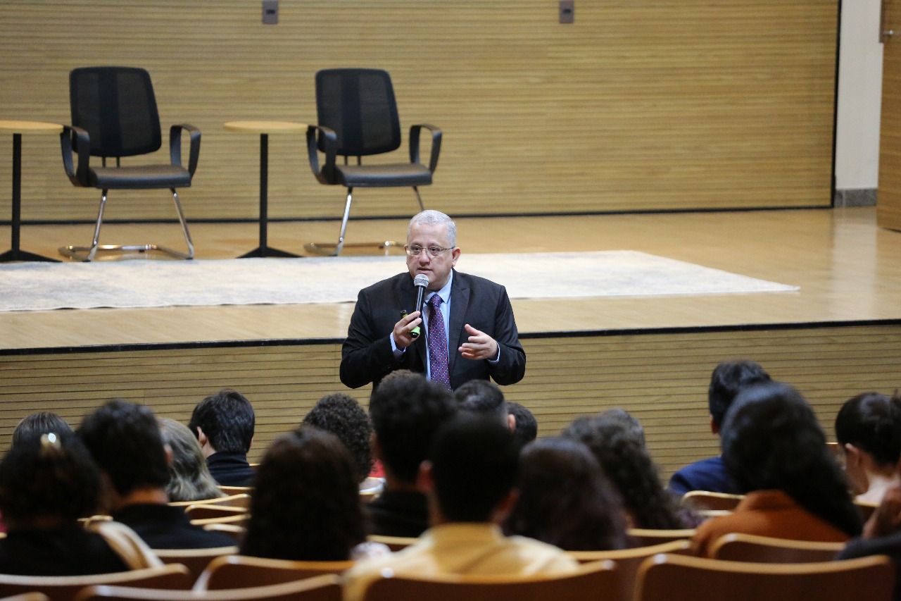Um homem fala ao público em um auditório. Ele segura um microfone e faz um gesto com a mão enquanto se dirige às pessoas sentadas nas poltronas. No palco, atrás dele, há três cadeiras e duas mesas pequenas. O ambiente é bem iluminado e tem aparência moderna, com paredes de madeira clara.