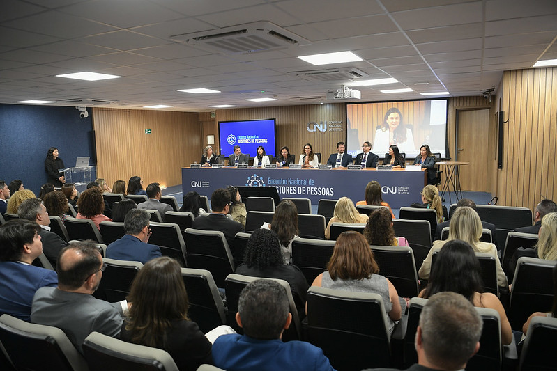 A foto mostra um auditório cheio, com participantes sentados voltados para uma mesa de autoridades posicionada na parte frontal da sala. Sobre o palco, cerca de dez representantes compõem a mesa do “Encontro Nacional de Gestores de Pessoas”, identificada por um painel azul com a identidade visual do CNJ. Ao fundo, dois telões exibem a transmissão do evento.