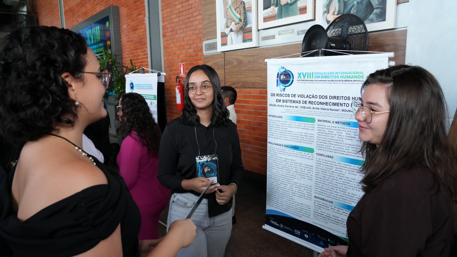 A imagem mostra três jovens conversando em frente a um pôster científico exposto no XVIII Congresso Internacional em Direitos Humanos. Duas delas, de frente para a câmera, usam crachá e estão posicionadas ao lado do painel intitulado “Os riscos de violação dos direitos humanos em sistemas de reconhecimento facial”.