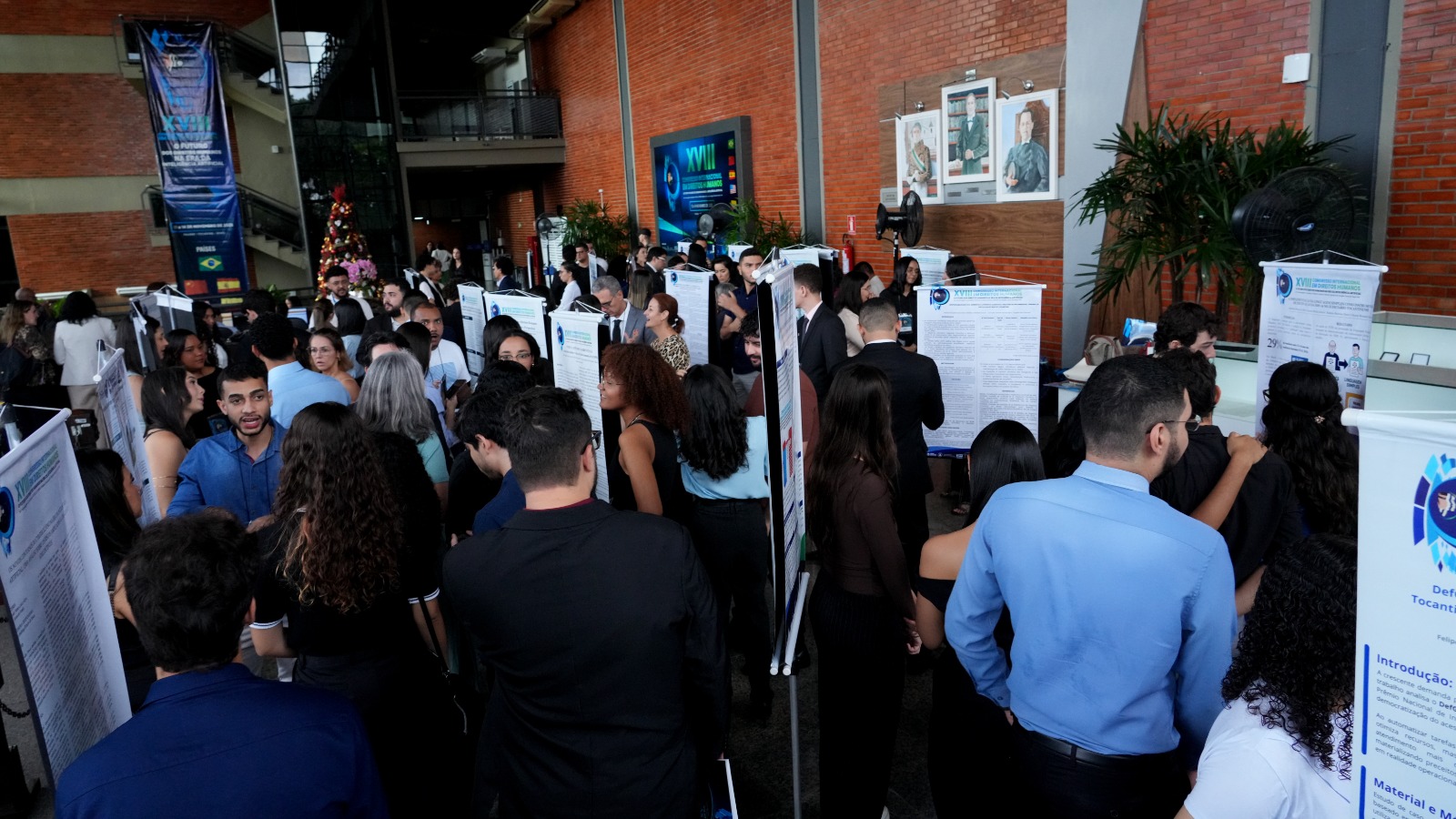 A foto mostra um grande hall movimentado, com dezenas de participantes circulando e apresentando pôsteres científicos alinhados em fileiras. As pessoas conversam em grupos diante dos painéis, enquanto o ambiente com paredes com revestimento cerâmico e banners do XVIII Congresso Internacional em Direitos Humanos compõem o cenário ao fundo.