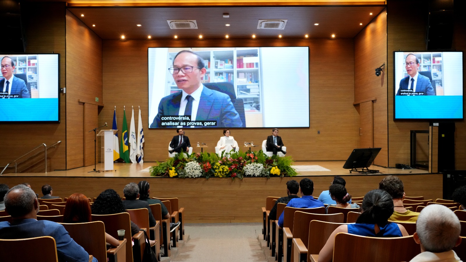 A foto mostra um auditório com público sentado assistindo a um painel no palco. Três participantes estão sentados em poltronas brancas diante de um arranjo de flores. No fundo, um telão exibe a imagem de um palestrante em videoconferência, também replicada em telas laterais. Bandeiras e um púlpito estão posicionados à esquerda do palco.