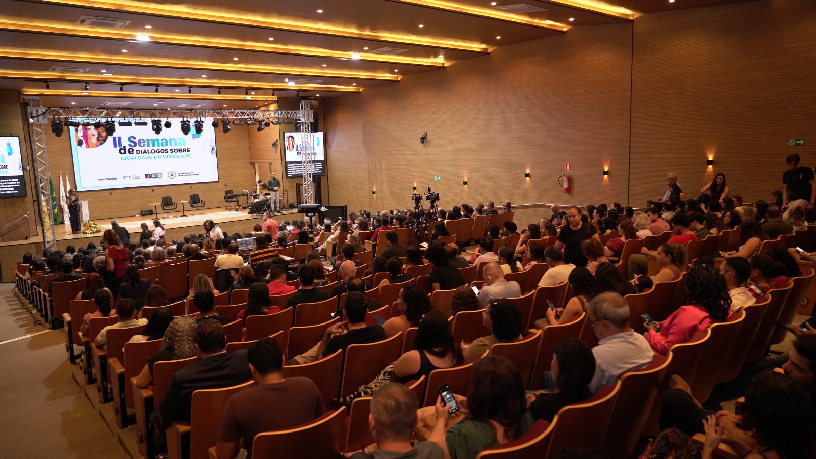 Foto do auditório do TJ lotado de pessoas com um painel no palco com a divulgação do evento