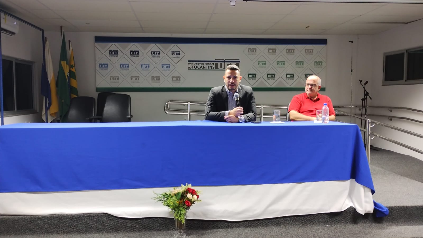 A foto mostra dois homens sentados à mesa principal de um auditório da UFT; um fala ao microfone e o outro está ao lado, com água sobre a mesa. Ao fundo, aparece o painel institucional e bandeiras.