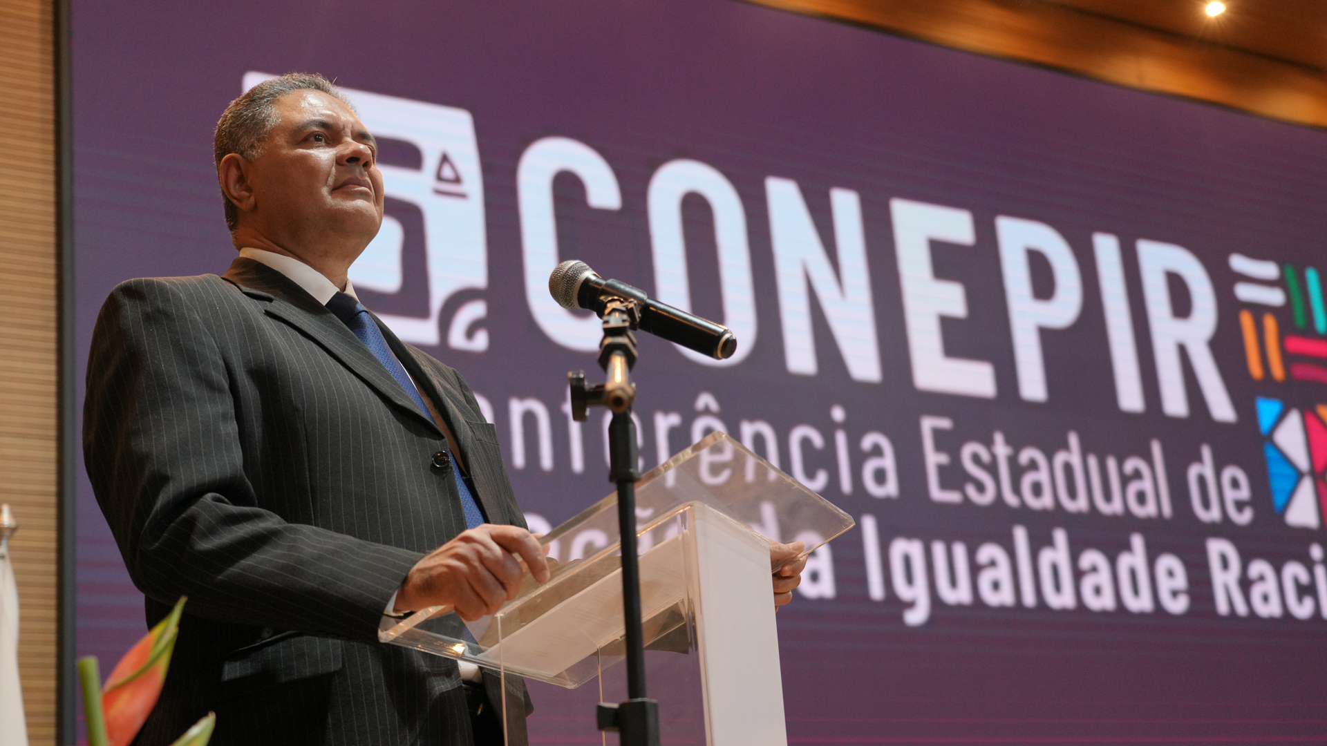 A foto mostra o juiz Rimabar em pé atrás de um púlpito transparente, falando ao microfone. Ele usa terno escuro e gravata azul. Ao fundo, há um telão com o nome “CONEPIR – Conferência Estadual de Promoção da Igualdade Racial”.