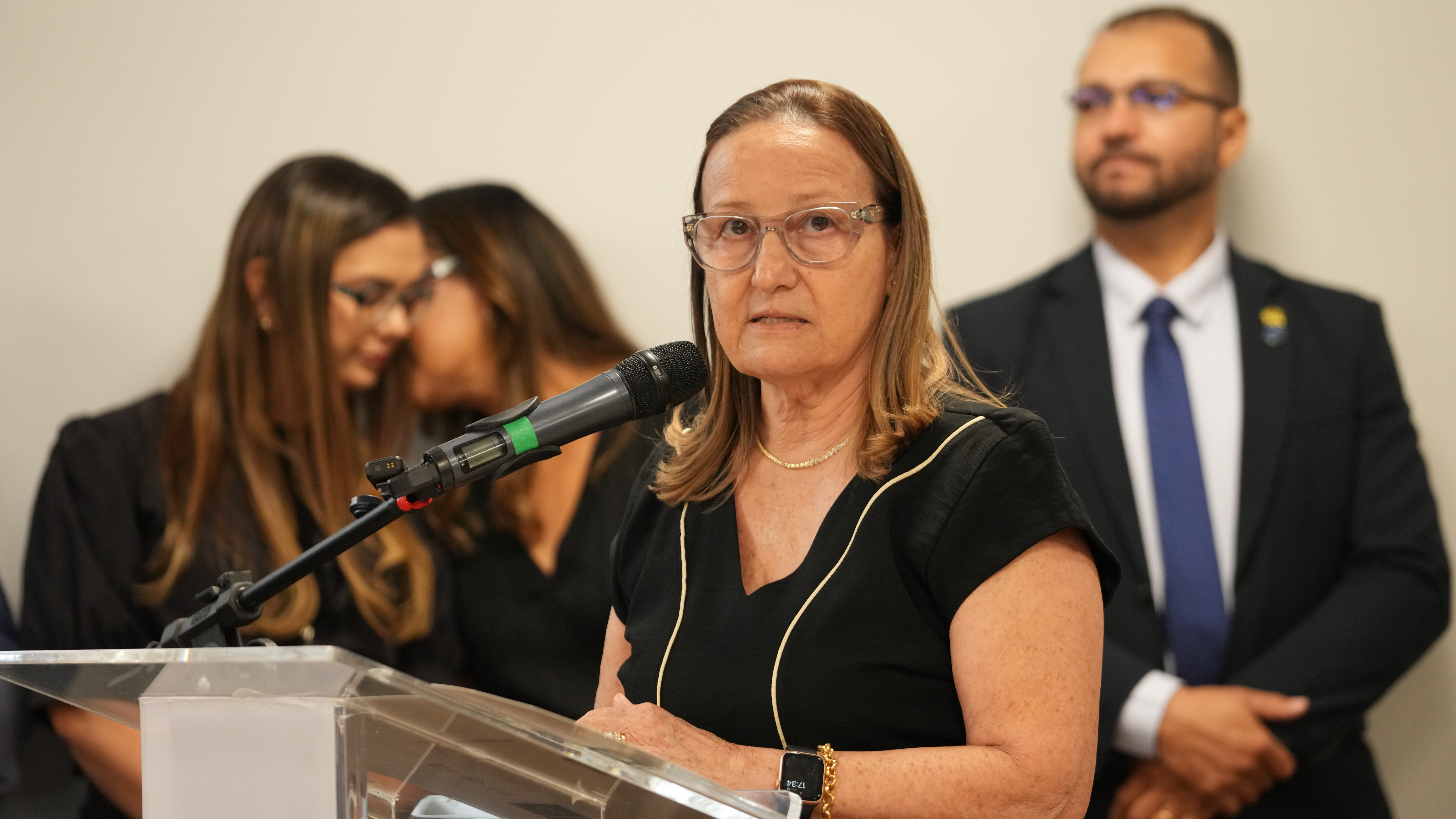 A foto mostra a desembargadora Maysa falando ao microfone em um púlpito transparente. Ela usa óculos e roupa preta. Ao fundo, duas mulheres conversam e um homem de terno observa a cena.