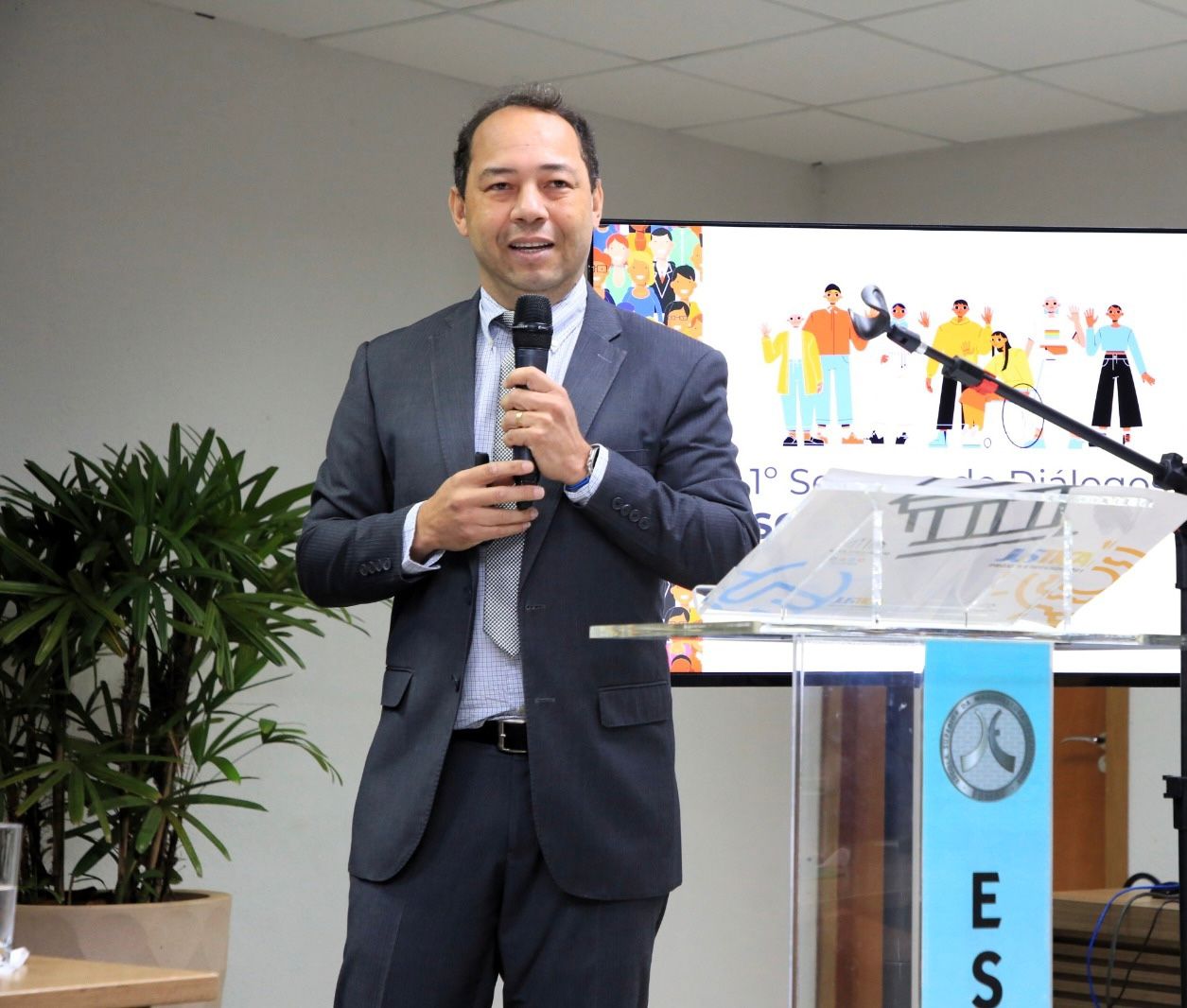 A foto mostra o juiz Jean,  de terno, falando ao microfone em um auditório. À frente dele há um púlpito transparente com identificação institucional. Atrás, uma tela exibe informações sobre um evento. À direita, um homem está sentado diante de um notebook.