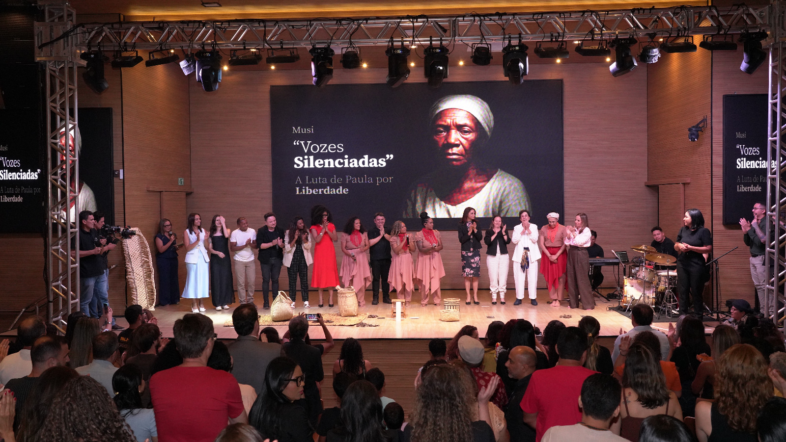 Público lota auditório enquanto o elenco de “Vozes Silenciadas – A luta de Paula por liberdade” está alinhado no palco, recebendo aplausos. Ao fundo, um grande painel exibe a imagem de uma mulher negra com expressão séria, em referência à personagem Paula. Luzes de espetáculo iluminam o cenário e músicos estão posicionados à direita do palco.