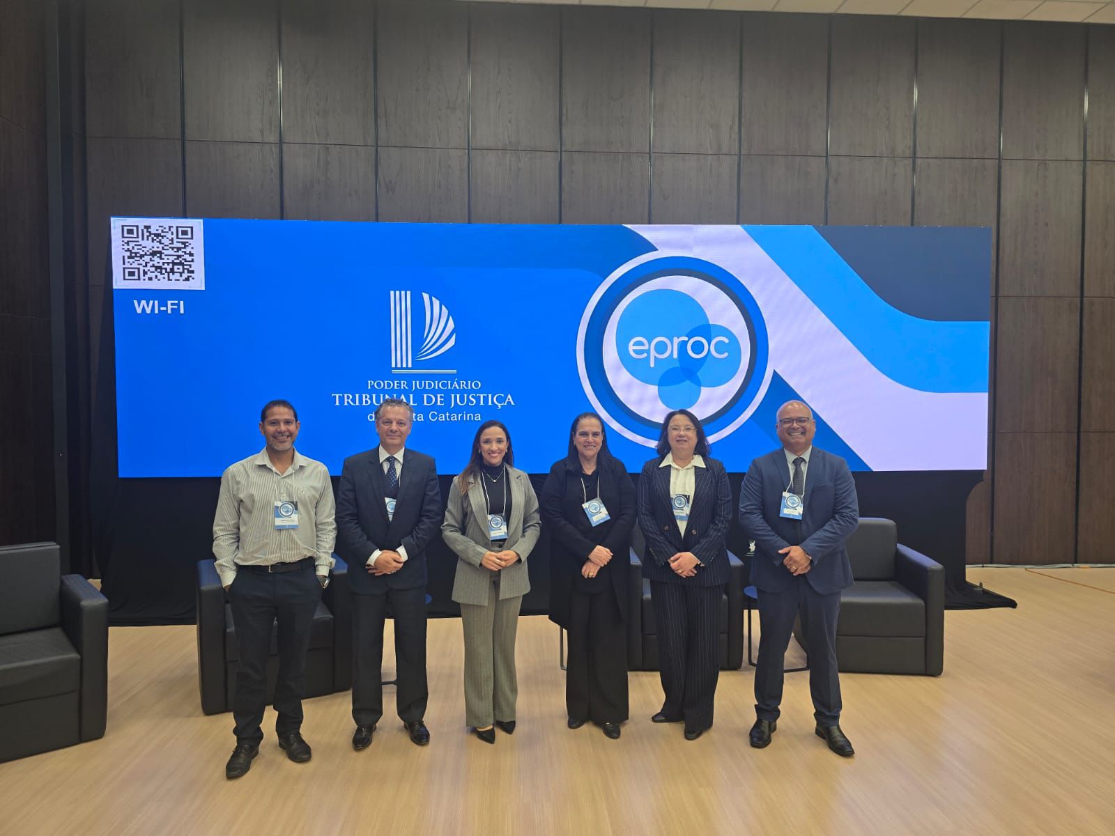 Seis pessoas posam para a foto em um auditório, todas usando crachá do evento. Elas estão alinhadas uma ao lado da outra, sorrindo, em frente a um grande painel azul com a marca do sistema eproc e o logotipo do Tribunal de Justiça de Santa Catarina