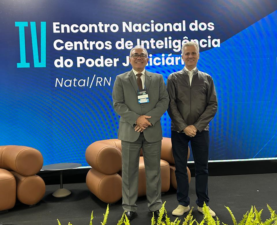 Desembargador Amaro e juiz Esmar posam lado a lado em frente a um painel azul com o texto “IV Encontro Nacional dos Centros de Inteligência do Poder Judiciário – Natal/RN”. À esquerda, o desembargador veste terno cinza e crachá; à direita, o juiz usa jaqueta marrom e calça azul escura. Atrás deles, há poltronas marrons e, à frente, um conjunto de plantas decorativas.