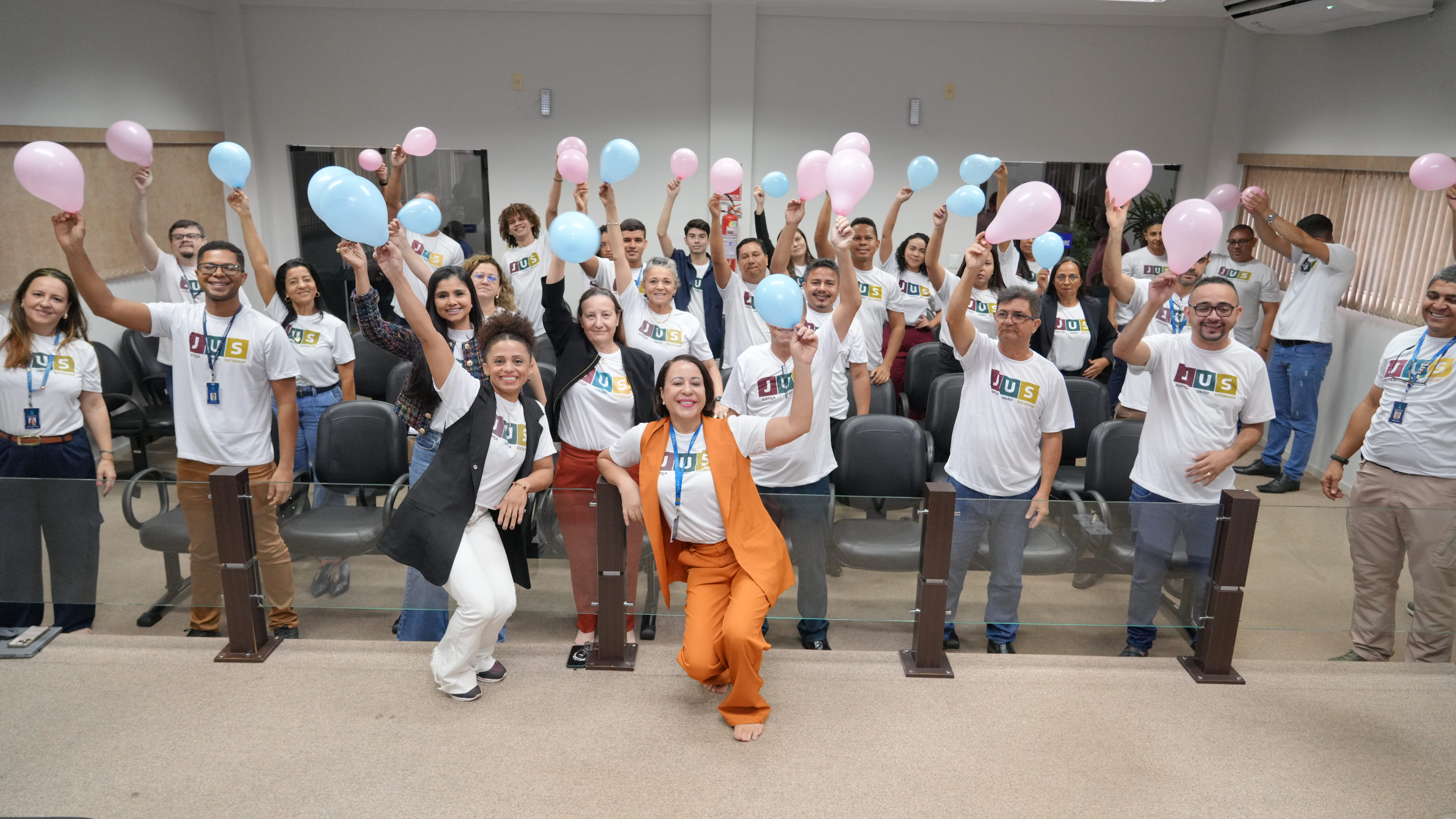 A imagem mostra um grupo numeroso de pessoas reunidas em um auditório, celebrando com entusiasmo. Todos vestem camisetas brancas com a palavra “JUS” estampada em letras coloridas e seguram balões rosa ou azul erguidos para o alto.  Na parte da frente, três mulheres posam sorrindo e em posição de destaque — uma delas, ao centro, está agachada e veste conjunto laranja sobre a camiseta. As demais pessoas, distribuídas pelas fileiras de cadeiras, também sorriem e participam do gesto coletivo, criando um clima de integração e alegria.  O ambiente é iluminado, com paredes claras, cortinas bege e cadeiras pretas ao fundo, transmitindo a ideia de uma ação institucional voltada para engajamento e sensibilização.