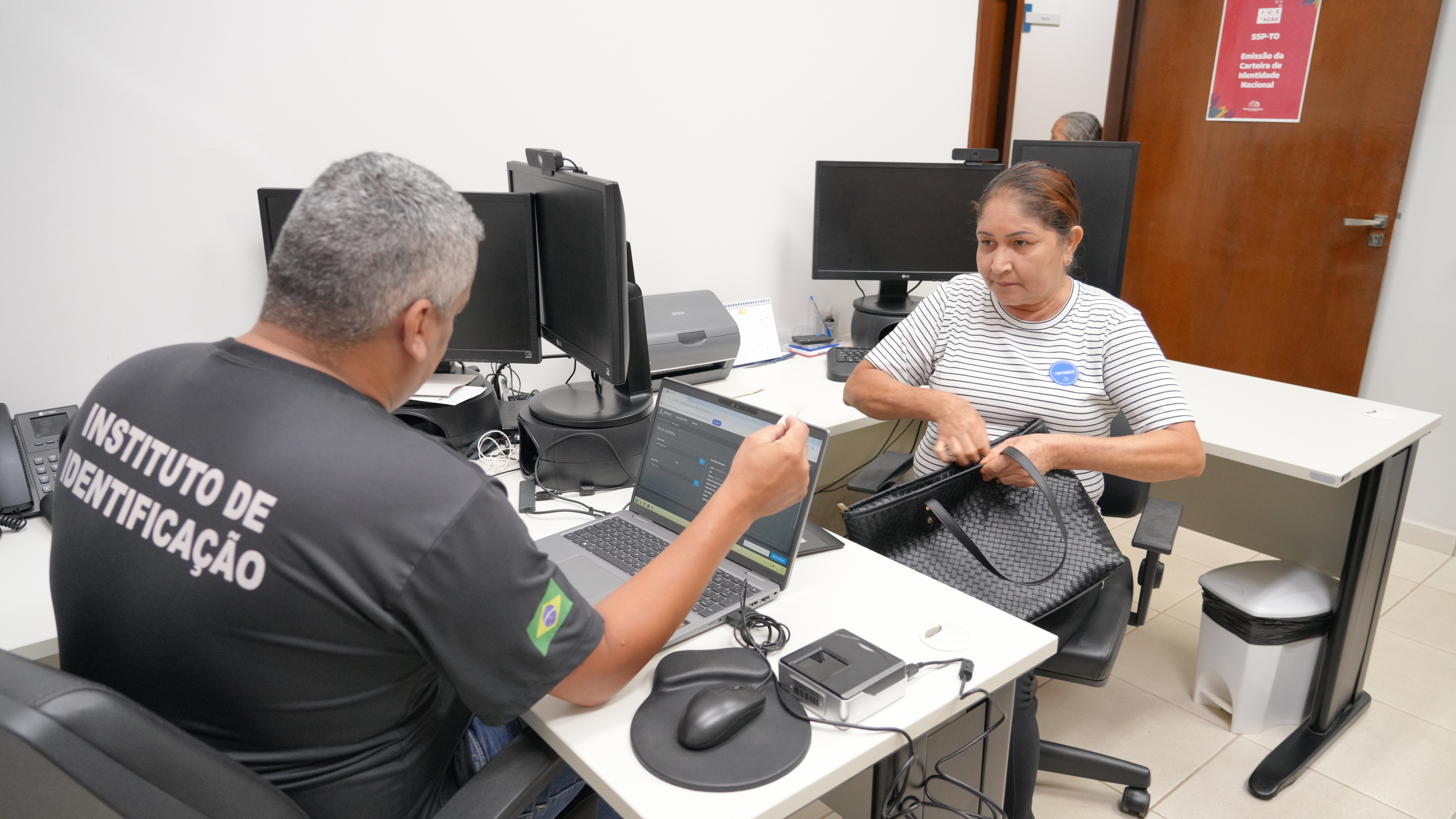 Na imagem, um servidor do Instituto de Identificação atende uma mulher em uma sala equipada com computadores. Ele está sentado diante de um notebook realizando um procedimento, enquanto ela, sentada à frente, mexe em uma bolsa preta.