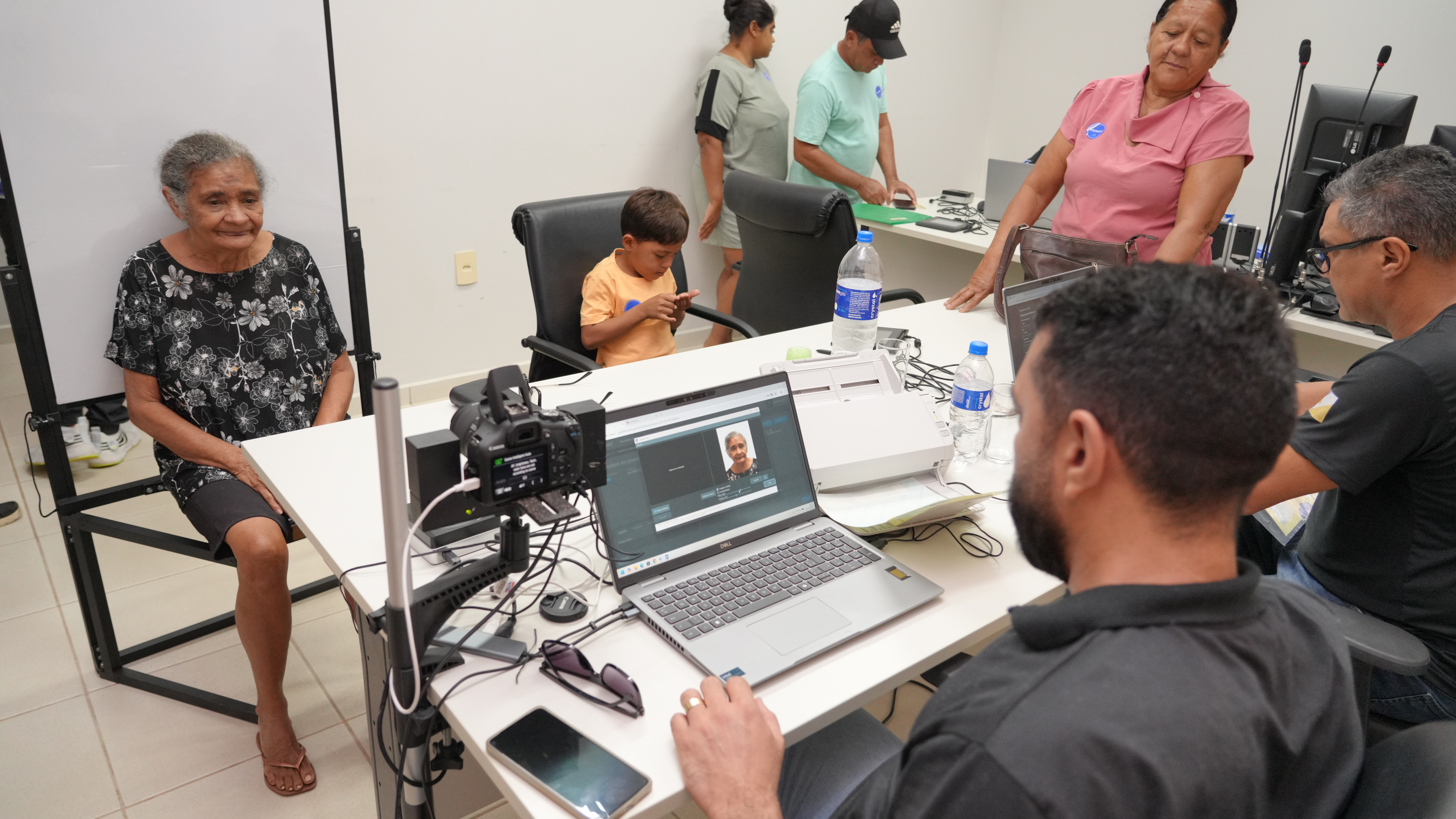 A foto mostra uma idosa sentada para registro fotográfico, enquanto um atendente opera um computador. Um menino está sentado ao lado mexendo no celular, e três pessoas aguardam ao fundo.