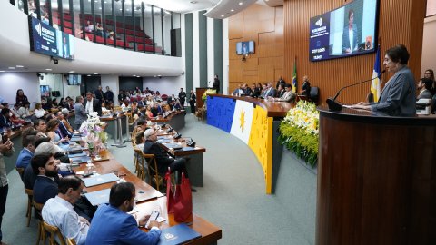 A imagem mostra o plenário da Assembleia Legislativa do Tocantins durante a sessão solene. A desembargadora Angela Issa Haonat aparece à direita, em pé na tribuna, fazendo seu discurso. O auditório está lotado, com autoridades, convidados e servidores sentados nas bancadas.