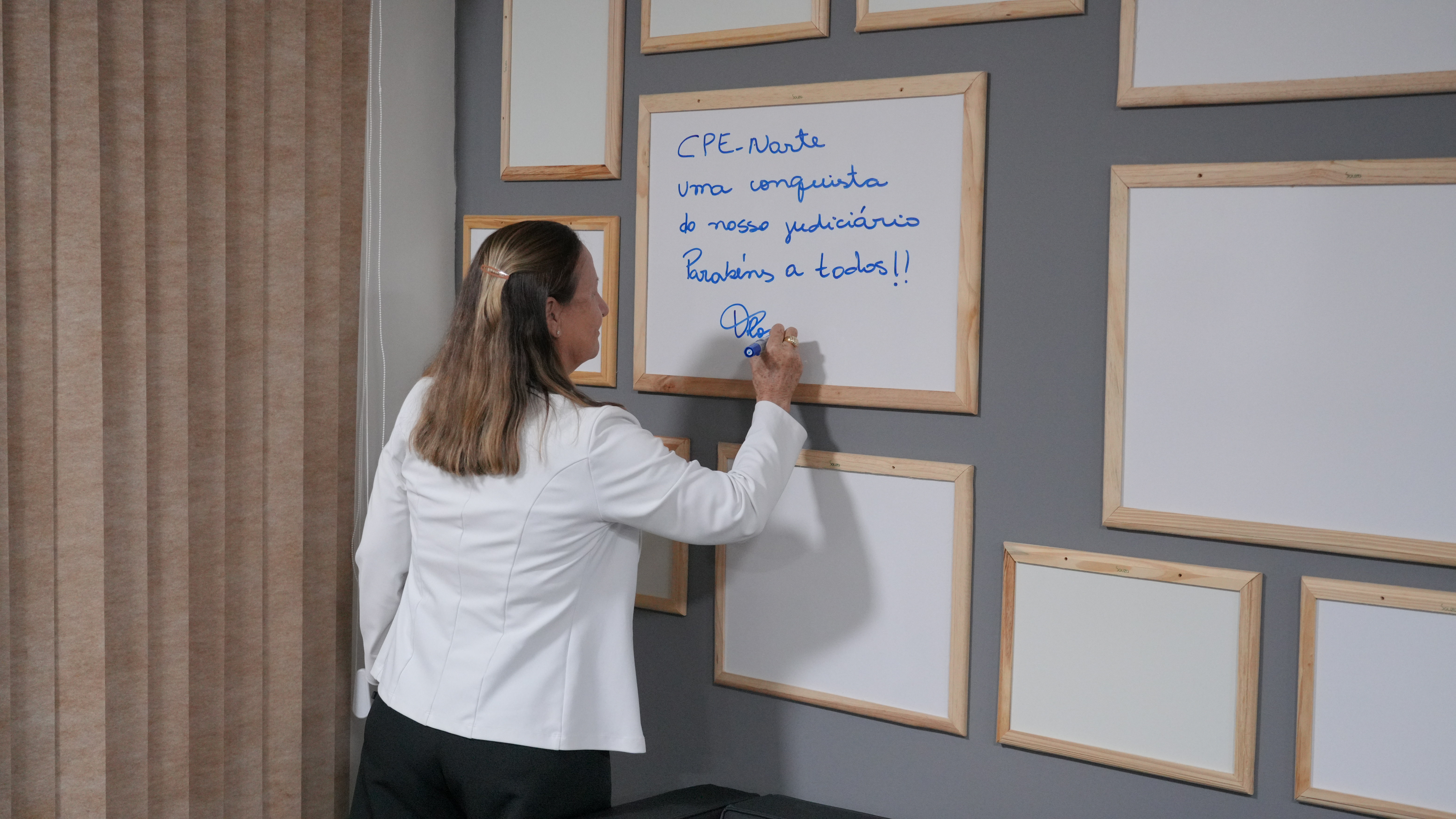 Uma mulher, vestindo blazer branco e calça escura, escreve uma mensagem em um quadro branco instalado em uma parede decorada com diversos quadros vazios de moldura clara. Ela está de costas para a câmera, segurando uma caneta azul enquanto finaliza o texto.  No quadro, lê-se: “CPE Norte Uma conquista do nosso Judiciário Parabéns a todos!!”