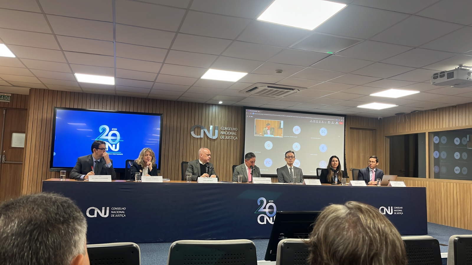 A imagem mostra uma mesa de autoridades durante um evento no Conselho Nacional de Justiça (CNJ). O ambiente é uma sala ampla, moderna, com paredes revestidas em madeira clara e teto com iluminação branca distribuída em painéis retangulares.  Ao centro, há uma mesa comprida coberta com um forro azul-marinho com o logotipo do CNJ e a marca de 20 anos. Sentados lado a lado, estão sete participantes, todos vestidos de forma formal — ternos e roupas sociais. Eles estão posicionados frente ao público, cada um com um microfone e copos de água à disposição.