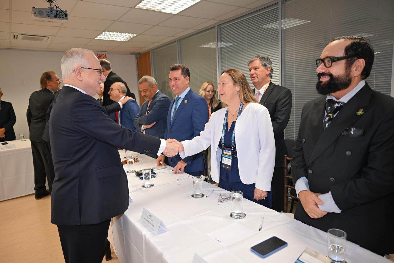 A foto mostra uma reunião formal, realizada em sala ampla e bem iluminada. No centro da cena, uma mulher vestindo blazer branco cumprimenta, com um aperto de mão, um homem de terno escuro e cabelos grisalhos. Ambos sorriem cordialmente, demonstrando respeito institucional.  Ao redor da mesa, outros participantes — homens e mulheres, todos trajando roupas formais — acompanham o momento. Alguns observam a saudação, outros conversam entre si. Sobre a mesa coberta com toalha branca há copos de água, canetas e placas de identificação.