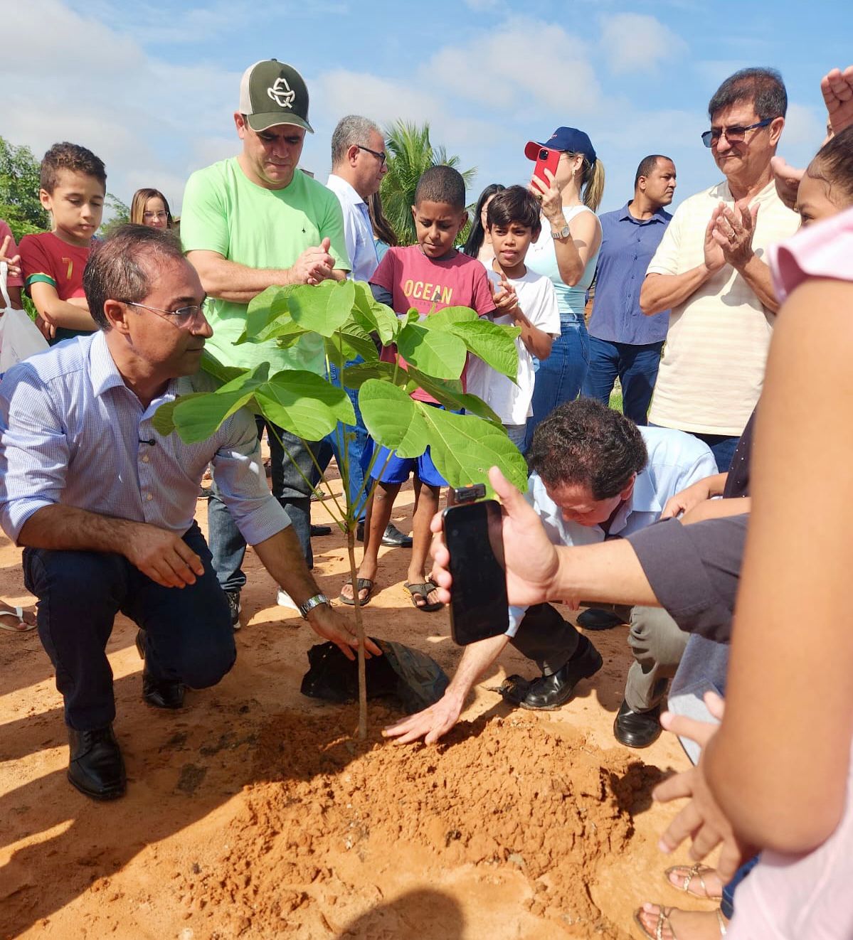 A foto mostra um grupo de pessoas durante o plantio de uma muda. Dois homens ajoelhados posicionam a planta no solo enquanto crianças e adultos observam ao redor. Alguns participantes batem palmas; outros registram a cena com o celular. A ação ocorre em área de terra batida sob céu aberto.