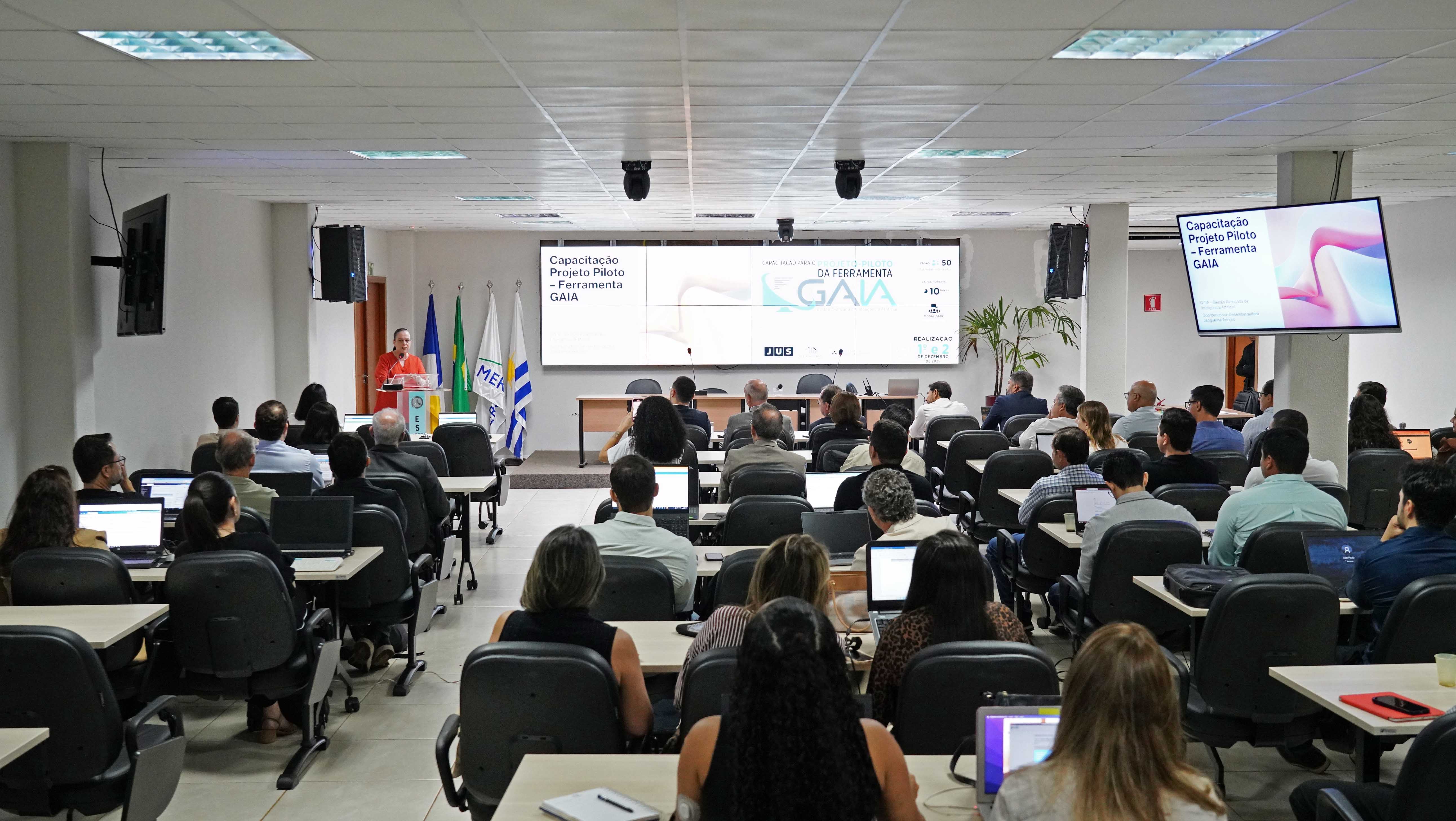 Auditório da Esmat lotado com magistrados(as) e servidores(as) sentados em carteiras, usando notebooks e acompanhando a capacitação da ferramenta GAIA, integrada ao eproc. Na parte da frente do ambiente, um palestrante está em pé, atrás de um púlpito, falando ao público. Ao fundo, há um grande telão com a apresentação “Capacitação Projeto Piloto – Ferramenta GAIA”, além das bandeiras do Brasil, Tocantins, TJTO e Esmat. O clima é de atenção e aprendizado sobre o uso de inteligência artificial no Judiciário.