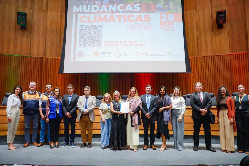 A foto mostra um grupo de pessoas enfileiradas no palco de um auditório, posando para a câmera. Ao fundo, há um telão com o título “Seminário Mudanças Climáticas 2025”. O grupo é composto por homens e mulheres, alguns em trajes formais e outros com uniforme de Defesa Civil. Todos estão de frente, em ambiente institucional.