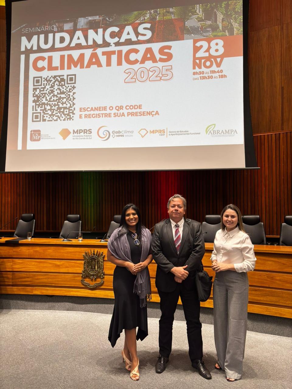A foto mostra três pessoas posando para a câmera no auditório onde ocorre o Seminário Mudanças Climáticas 2025. Elas estão em pé, à frente da mesa principal do plenário, que possui cadeiras pretas e painel de madeira ao fundo. Acima delas, um telão exibe o título do evento, a data e logomarcas das instituições organizadoras. As duas mulheres vestem roupas sociais, e o homem, traje formal com gravata. Todos estão de frente e sorrindo.
