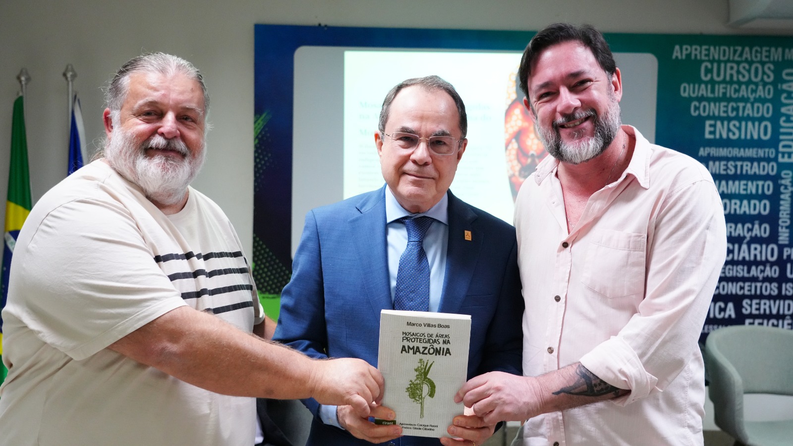 A imagem mostra três homens posando juntos e sorrindo, enquanto seguram o livro “Mosaicos de Áreas Protegidas na Amazônia”. Ao fundo tem bandeiras e um painel com palavras relacionadas a educação, cursos e formação.