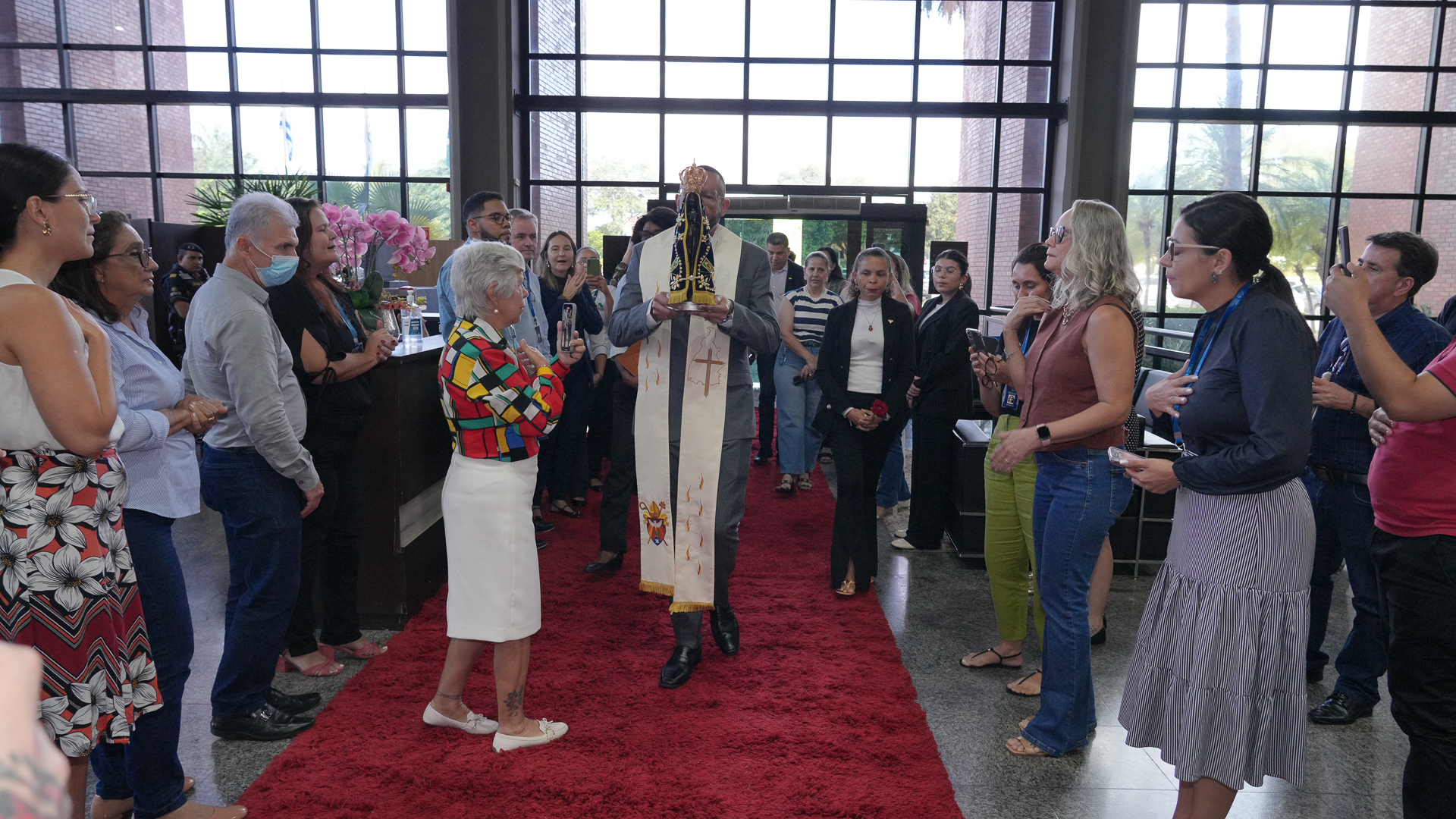 A fotografia mostra o momento de acolhimento da imagem  peregrina de Nossa Senhora Aparecida no hall de entrada do Tribunal de Justiça do Tocantins. No centro da cena, um padre caminha sobre um tapete vermelho segurando a Santa. Ao redor, há um grupo de pessoas — homens e mulheres de diferentes idades.