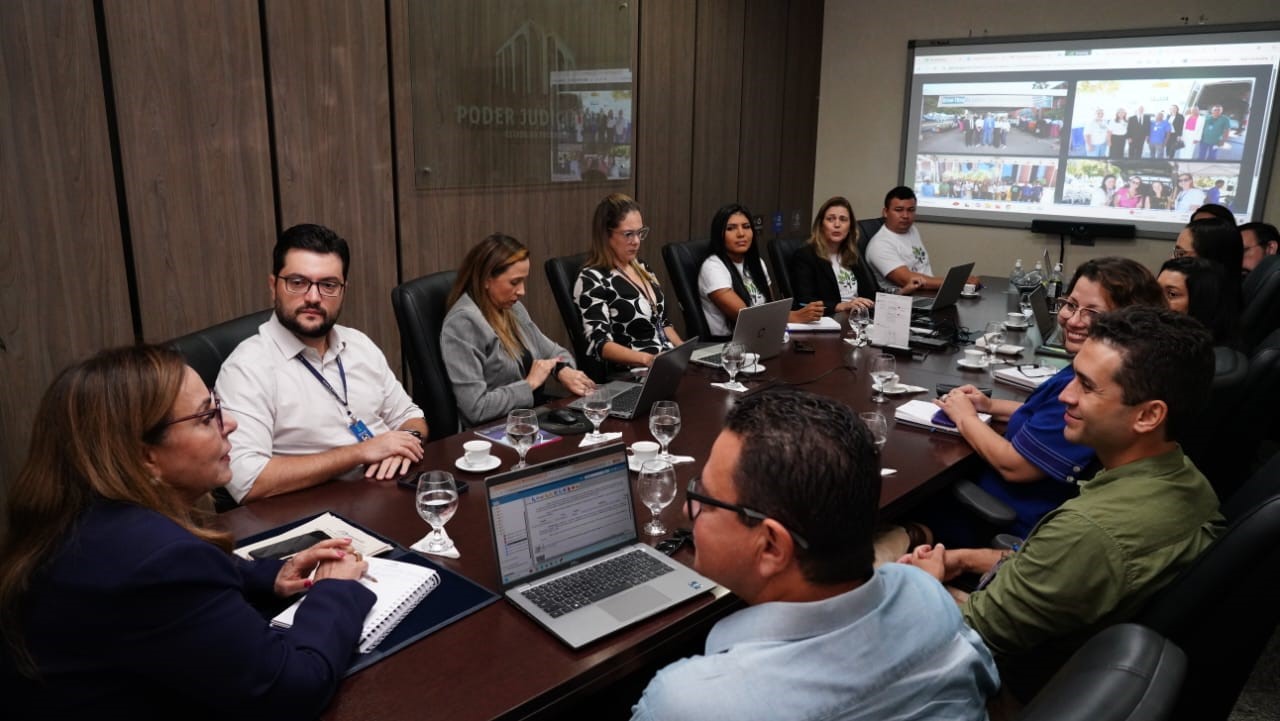 Um grupo de homens e mulheres está sentado ao redor de uma mesa retangular, em ambiente formal, dialogando. Ao centro, a desembargadora Ãngela Prudente,  uma mulher de pele clara que usa roupa azul marinho, conduz a conversa. Sobre a mesa, há copos com água, xícaras de café, blocos de anotações, celulares e um notebook. O clima é de diálogo e alinhamento, com todos atentos à discussão.