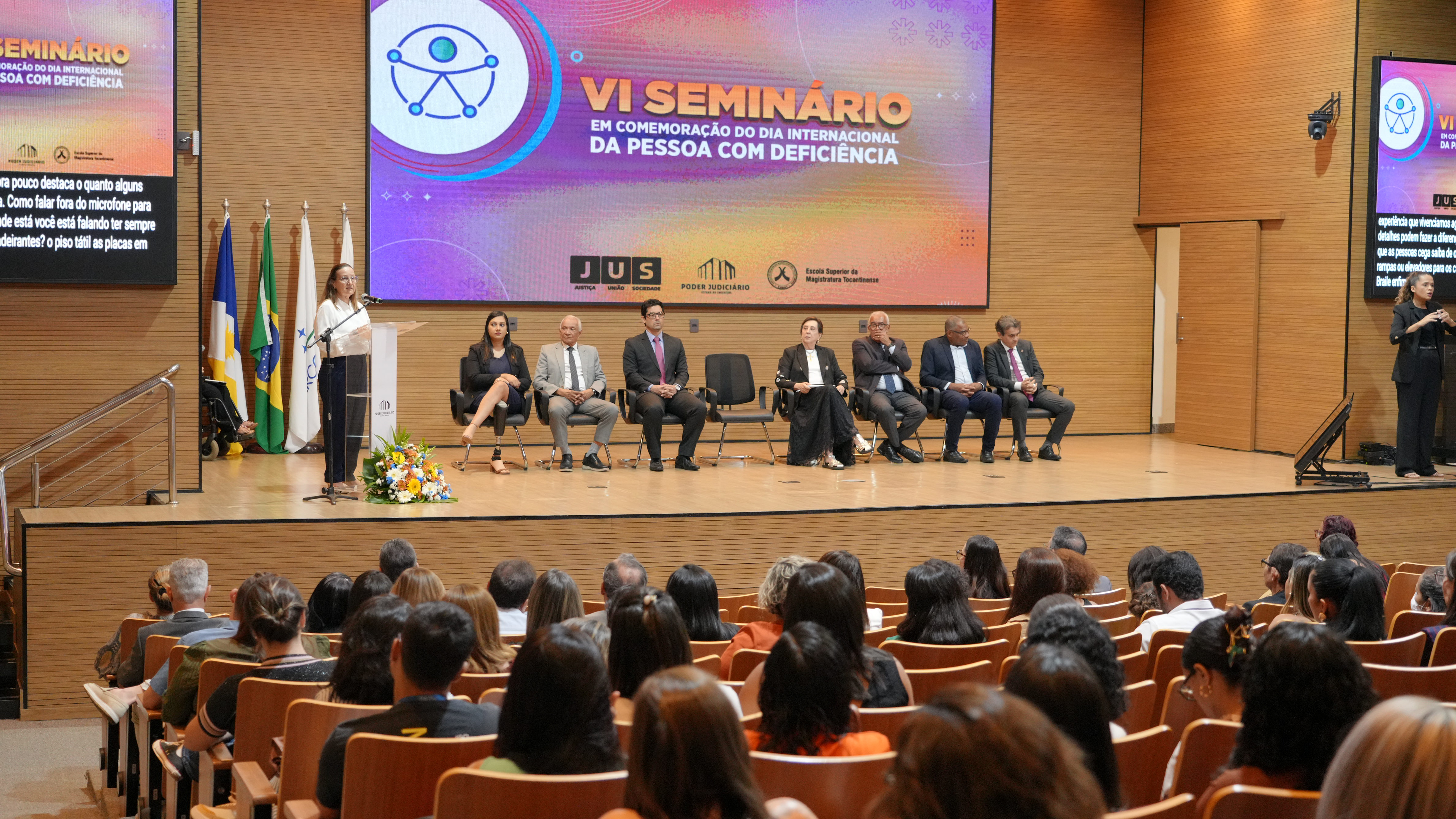 A imagem mostra um auditório cheio, com várias pessoas assistindo VI Seminário em Comemoração do Dia Internacional da Pessoa com Deficiência. No palco, há sete autoridades sentadas e a presidente do TJTO, desembargadora Maysa Vendramini Rosal fala ao microfone em um púlpito. À direita, uma intérprete de Libras traduz a fala. Um telão ao fundo exibe o título do evento.