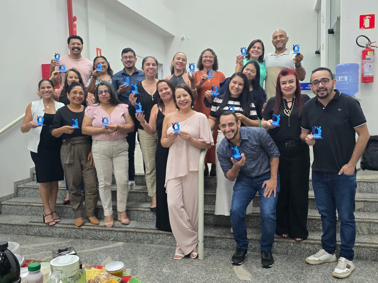 Grupo de servidores(as) posa para foto em ambiente interno do fórum, segurando troféus azuis do Programa Judiciário na Medida, em registro do encerramento das atividades.