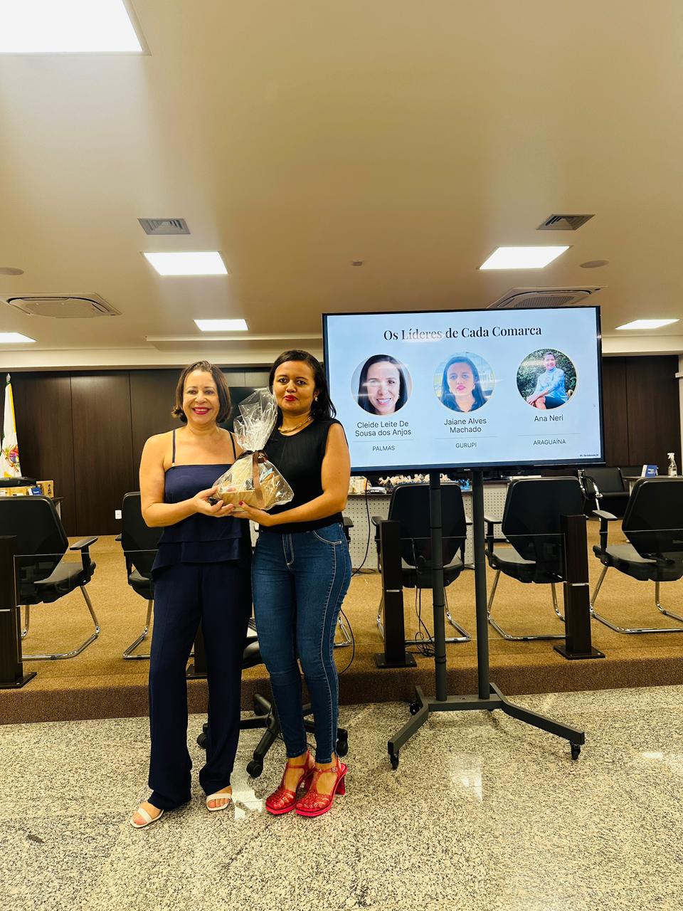 Sandra e a servidora Jaiane posam para foto em auditório durante o encerramento do Programa Judiciário na Medida 2025. A servidora, à direita, recebe um prêmio simbólico, enquanto, ao fundo, um telão exibe a identificação dos líderes das comarcas participantes.