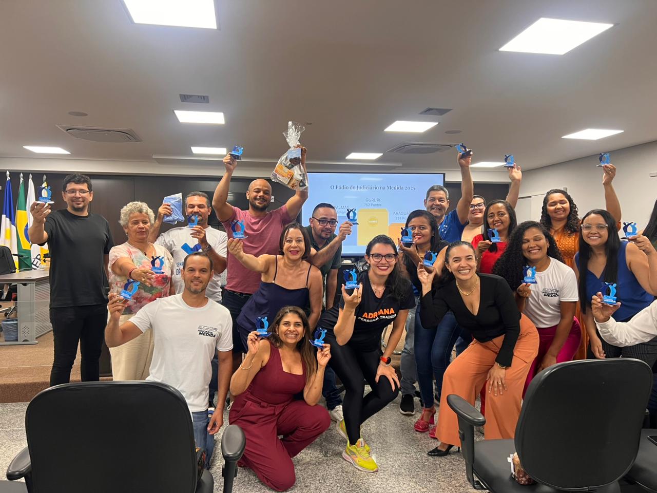 Grupo de servidores(as) participa do encerramento do Programa Judiciário na Medida 2025 em auditório, exibindo troféus e prêmios simbólicos, em registro coletivo de celebração e reconhecimento.